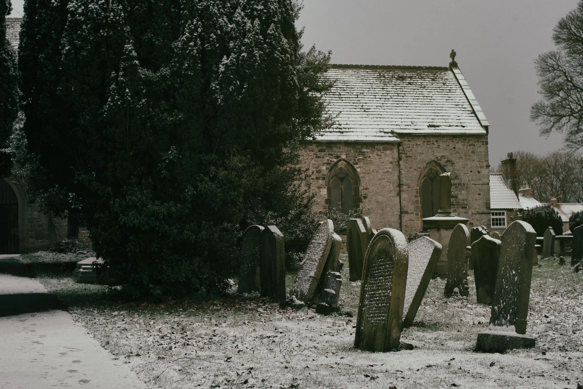 A snowy graveyard with tombstones covered in snow, a stone church with arched windows, and leafless trees in the background, during overcast winter weather.