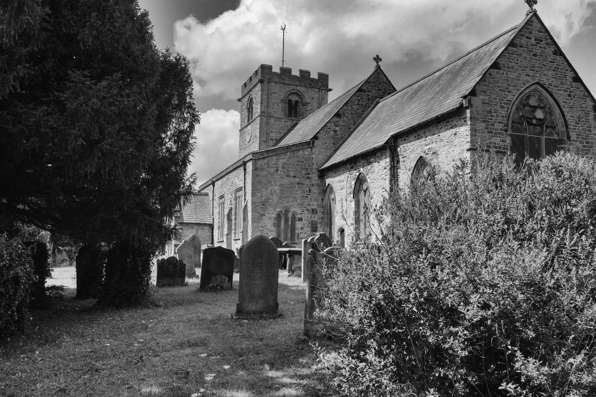 Black and white photo of St. Michael's Church with a tower, pointed roof, large arched window, and a graveyard with headstones in the foreground.