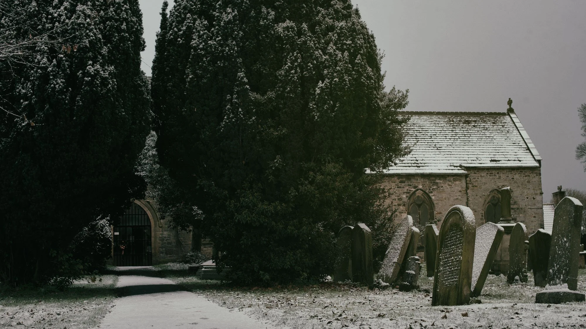 Snow-covered gravestones in a graveyard with a stone church building in the background, large trees also covered in snow, overcast sky