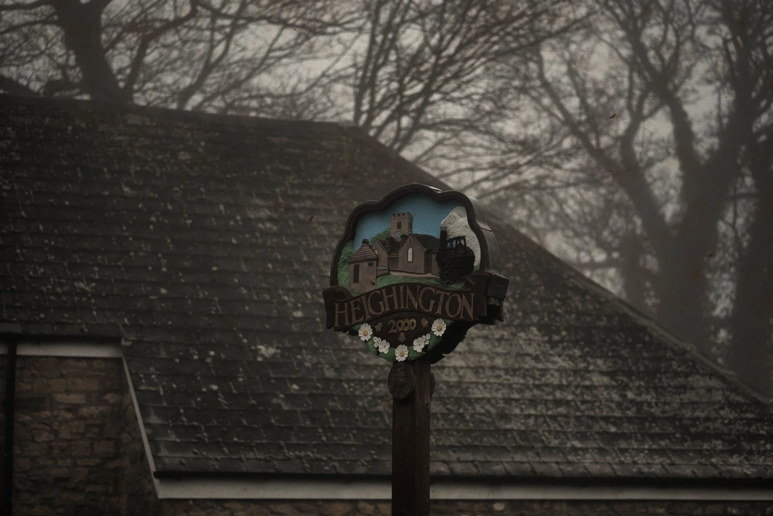 A hanging sign with the word 'Heington' and a depiction of a church, along with the year 1990, surrounded by white daisies, is in front of a house with a mossy shingled roof and leafless tree branches overhead on a cloudy day.
