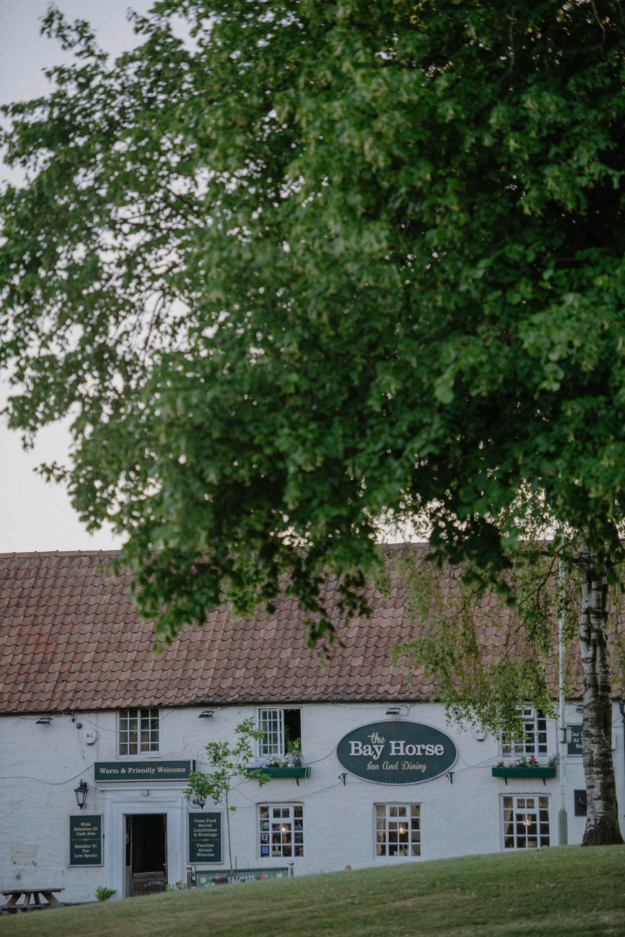 A white building with the sign 'the Bay Horse Inn And Dining' and additional signs advertising the menu and services, with a lawn and some small trees in front and a large leafy tree partially obscuring the view, with a tiled roof on the building.