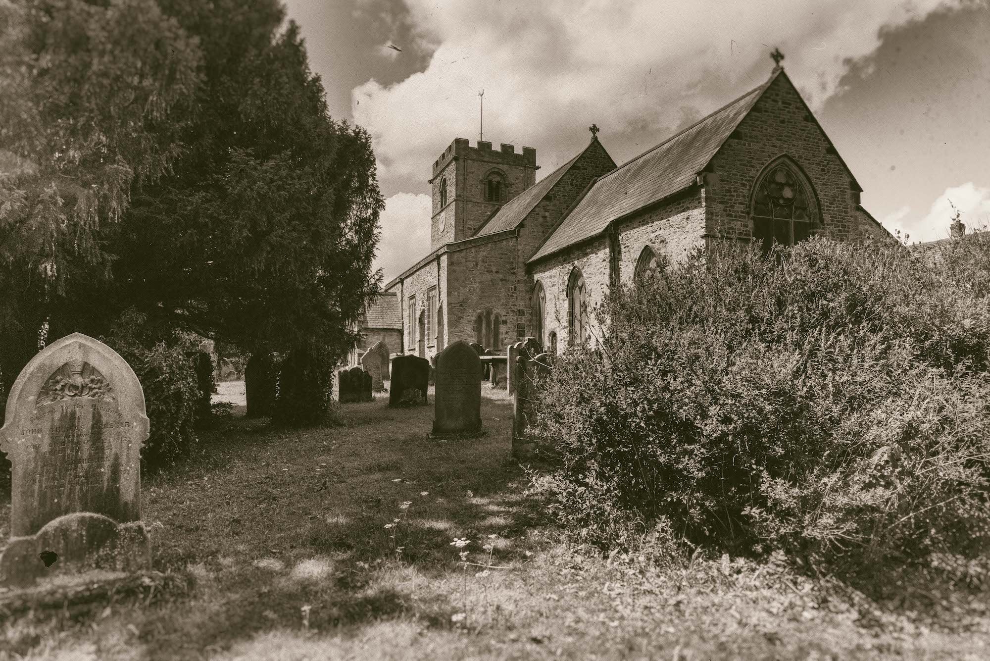 Black and white photo of an St. Michaels's church with a graveyard in front, headstones among trees and bushes, cloudy sky above.