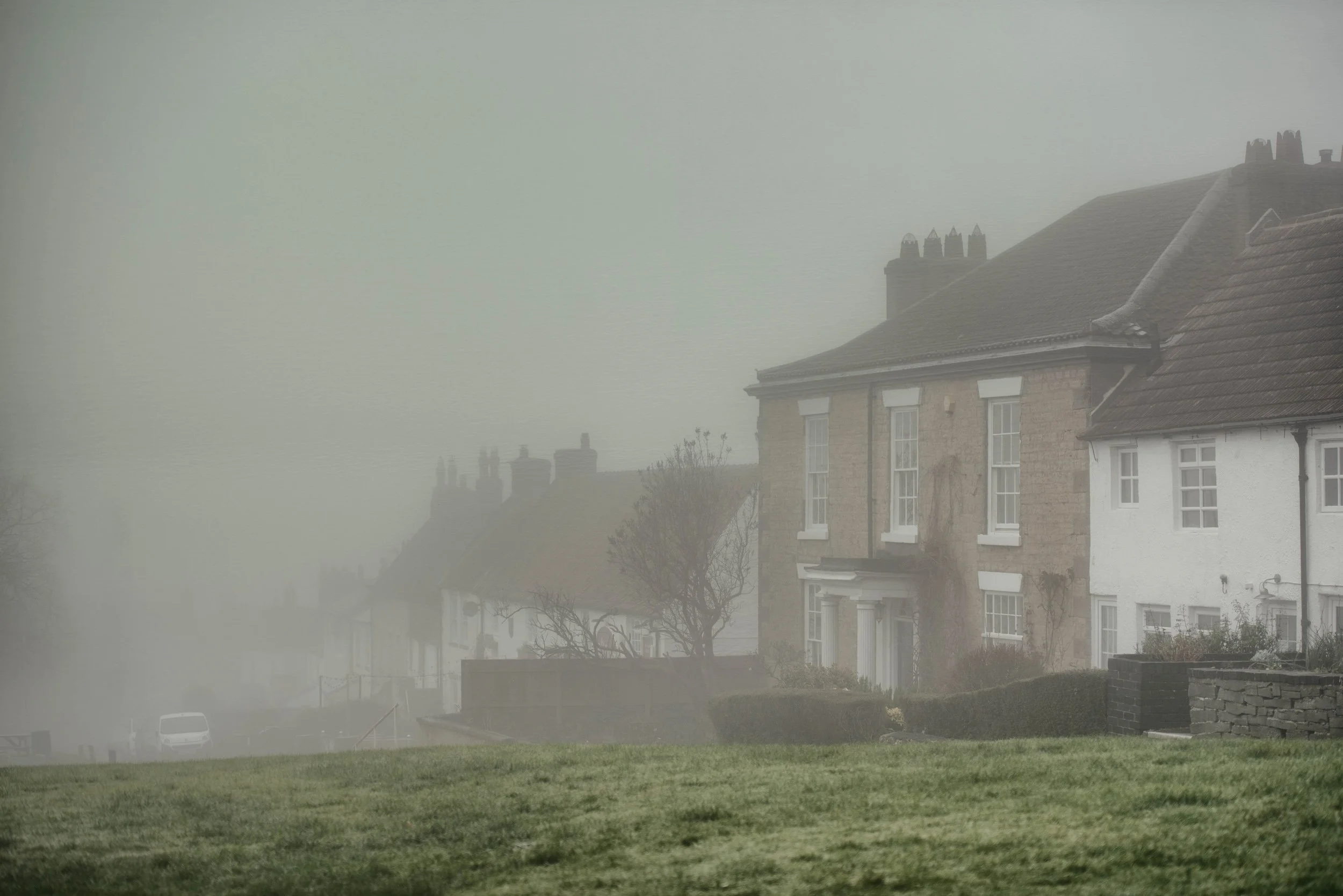 A foggy neighborhood street with houses, trees, and cars faintly visible in the background.