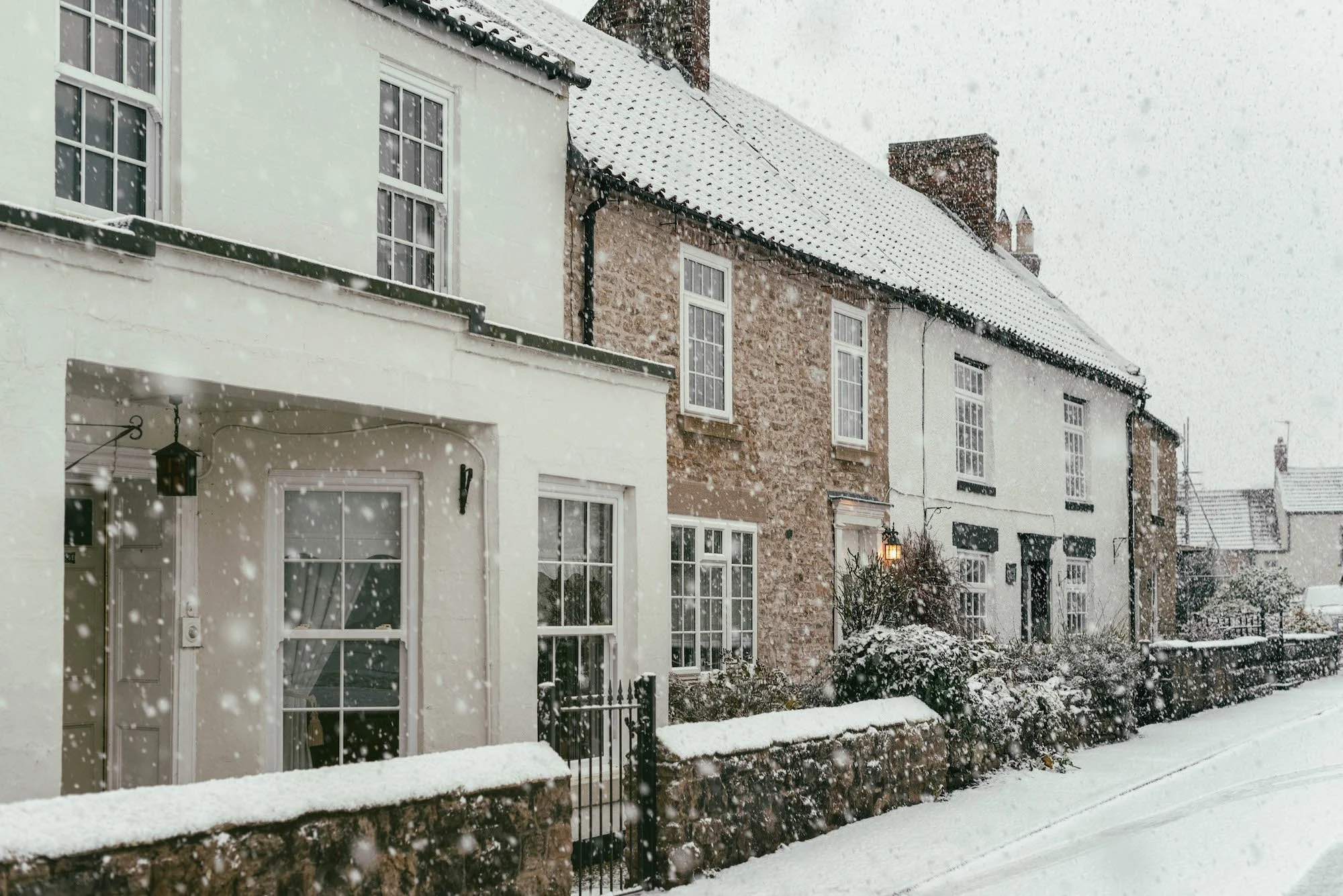 A row of houses with snow-covered rooftops and a snow-covered sidewalk in a quiet neighborhood during a snowfall.
