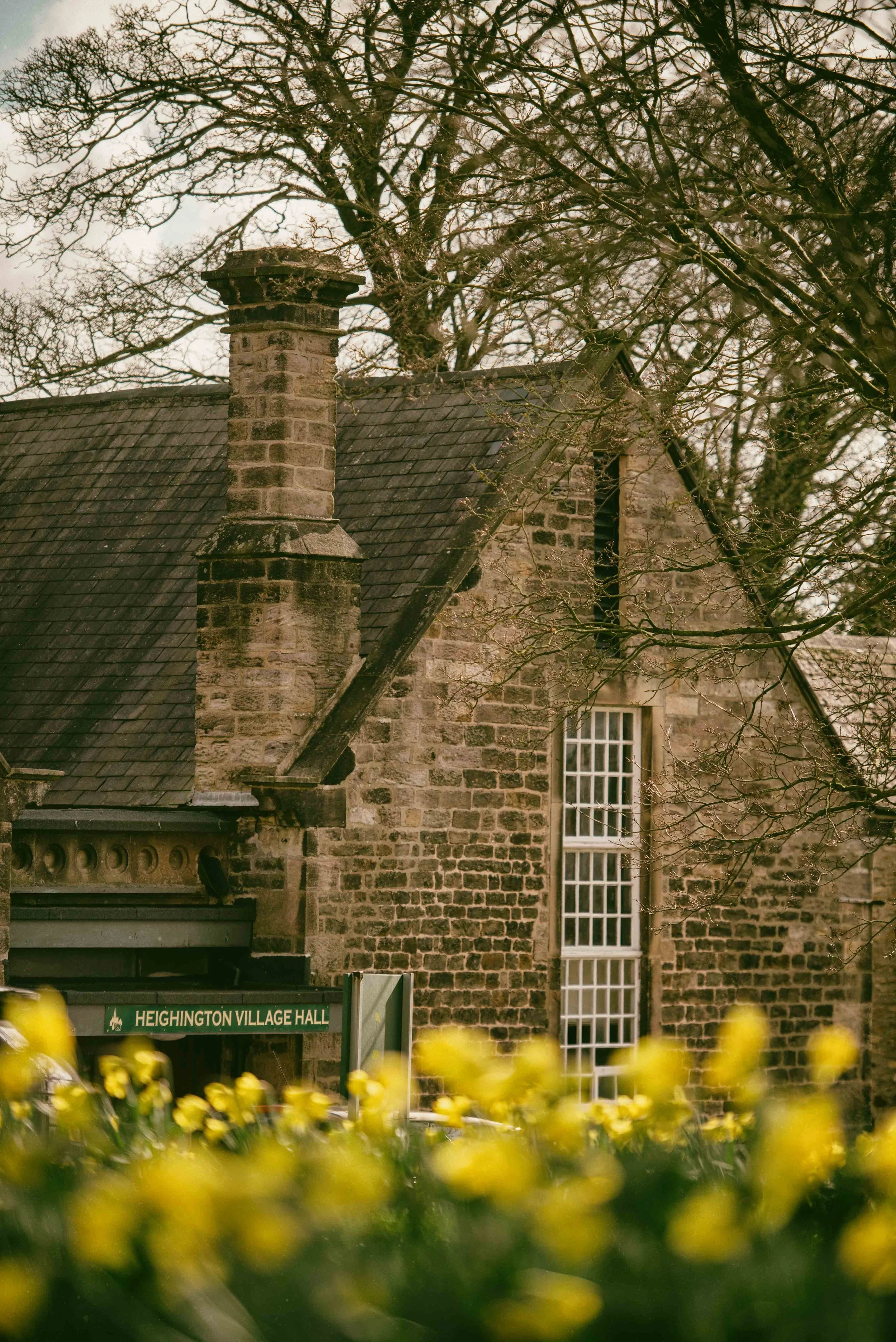 Stone building with a chimney and tall window, trees in the background, and a sign reading 'Heighington Village Hall' at the entrance.