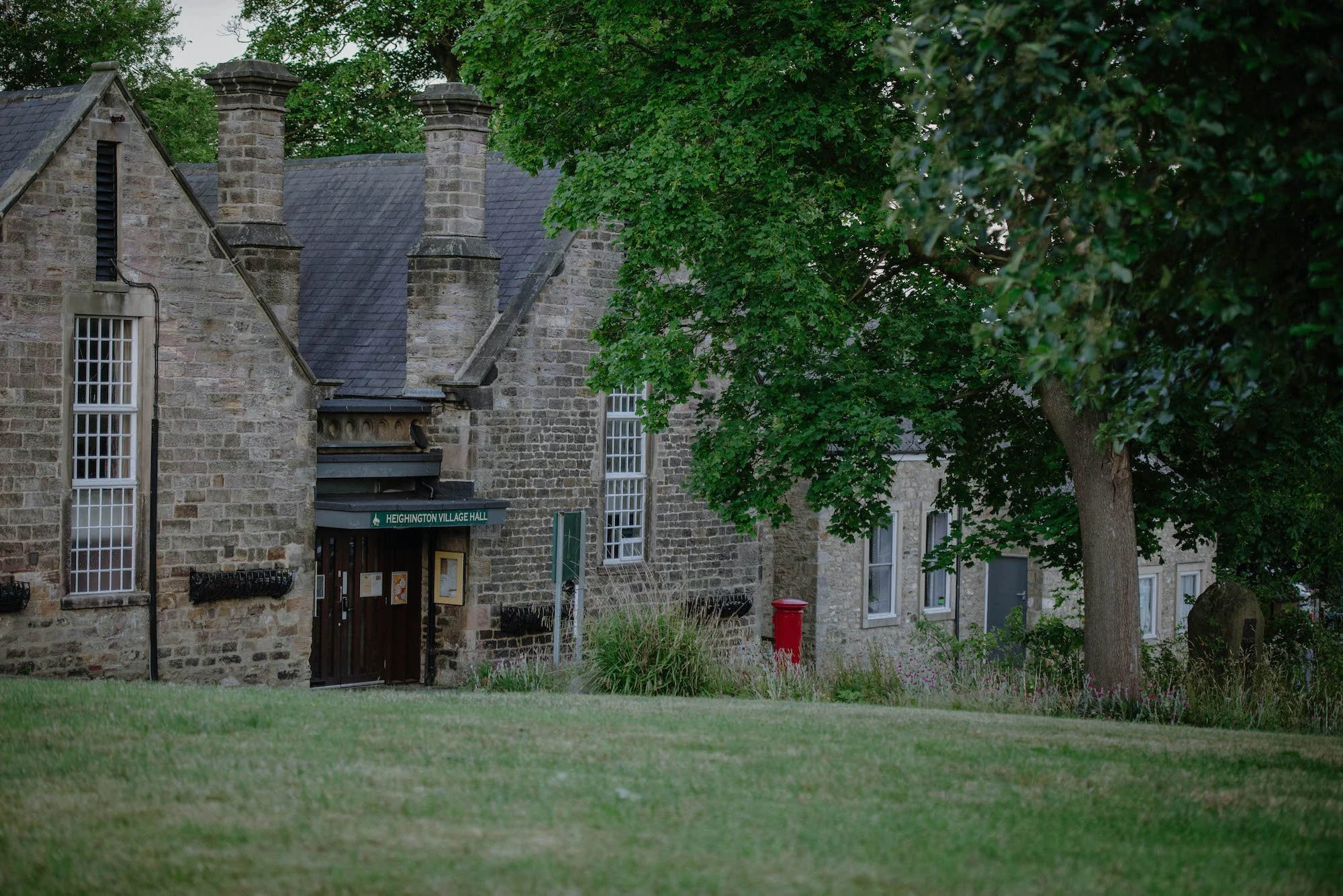A stone building with several chimneys, tall windows, and a green street sign that says "Heingham Village Hall". There are trees with lush green leaves and a well-maintained lawn in front of the building.