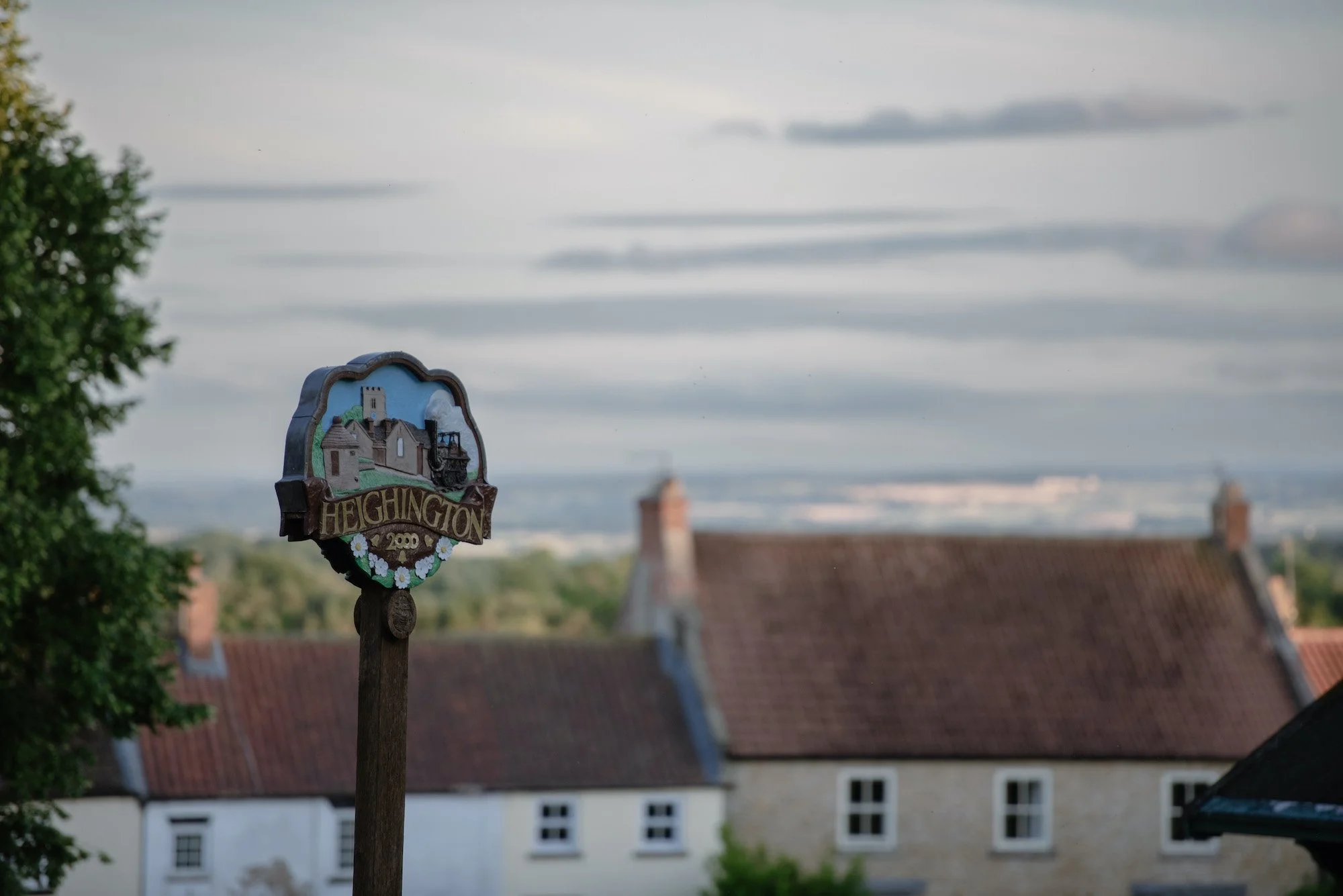 A decorative sign with the word "Heington" and an illustration of a small village, mounted on a wooden post, with a background of rooftops and a cloudy sky.