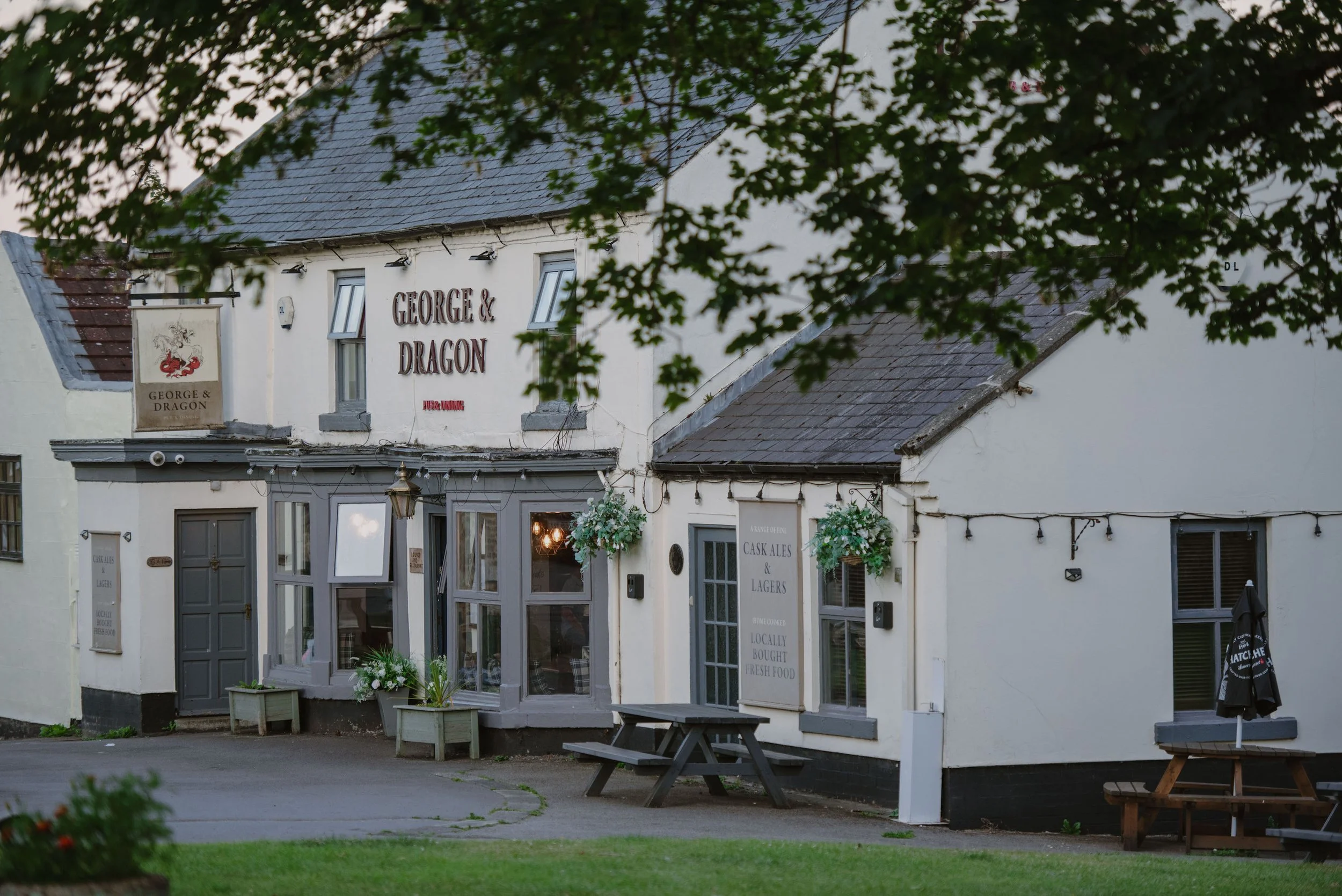 Exterior view of a pub named 'George & Dragon' with a white facade, gray trim, and string lights, surrounded by greenery and outdoor seating.