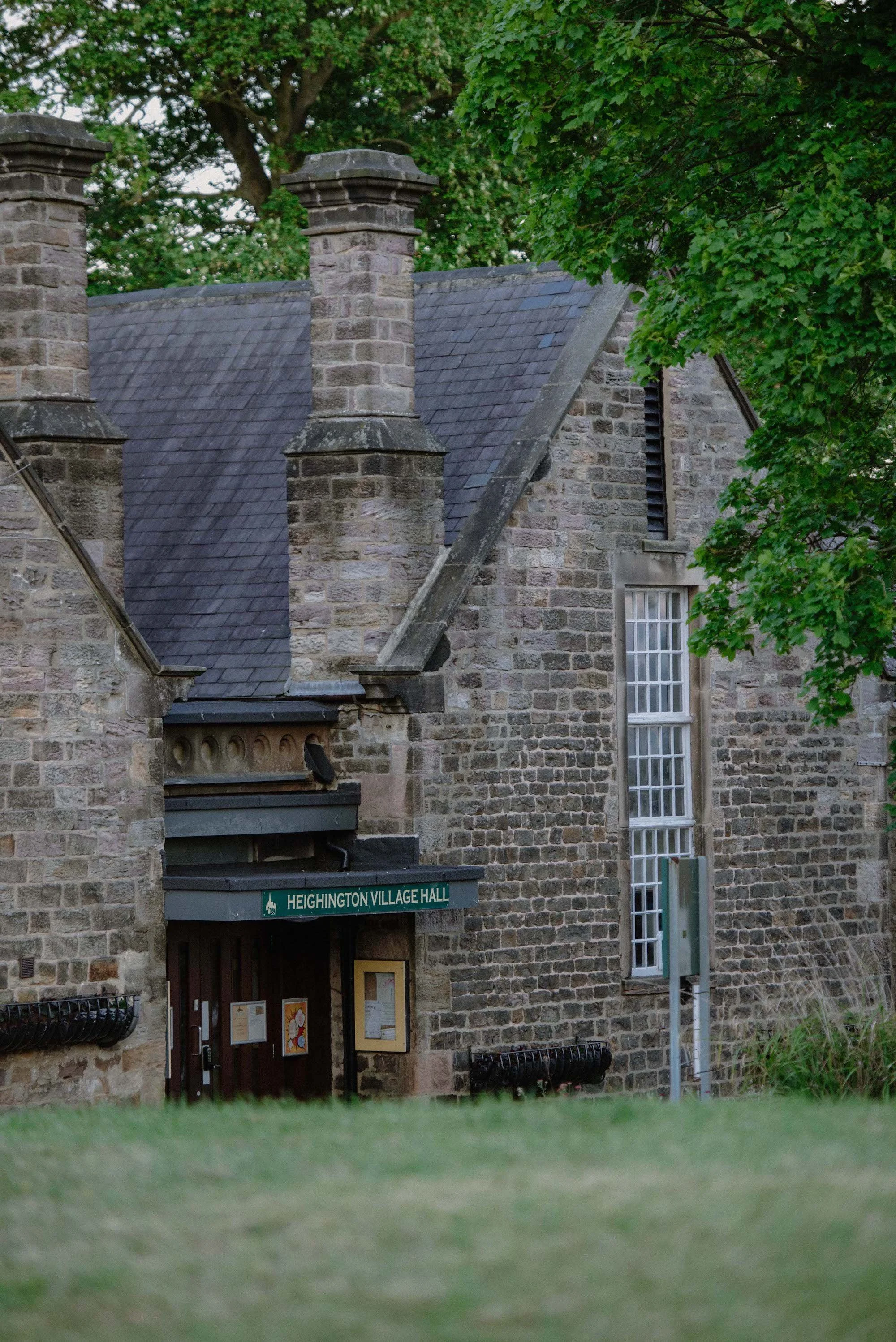 Stone building with a sign that reads "Heighington Village Hall" and a window below it, surrounded by green trees and grass.