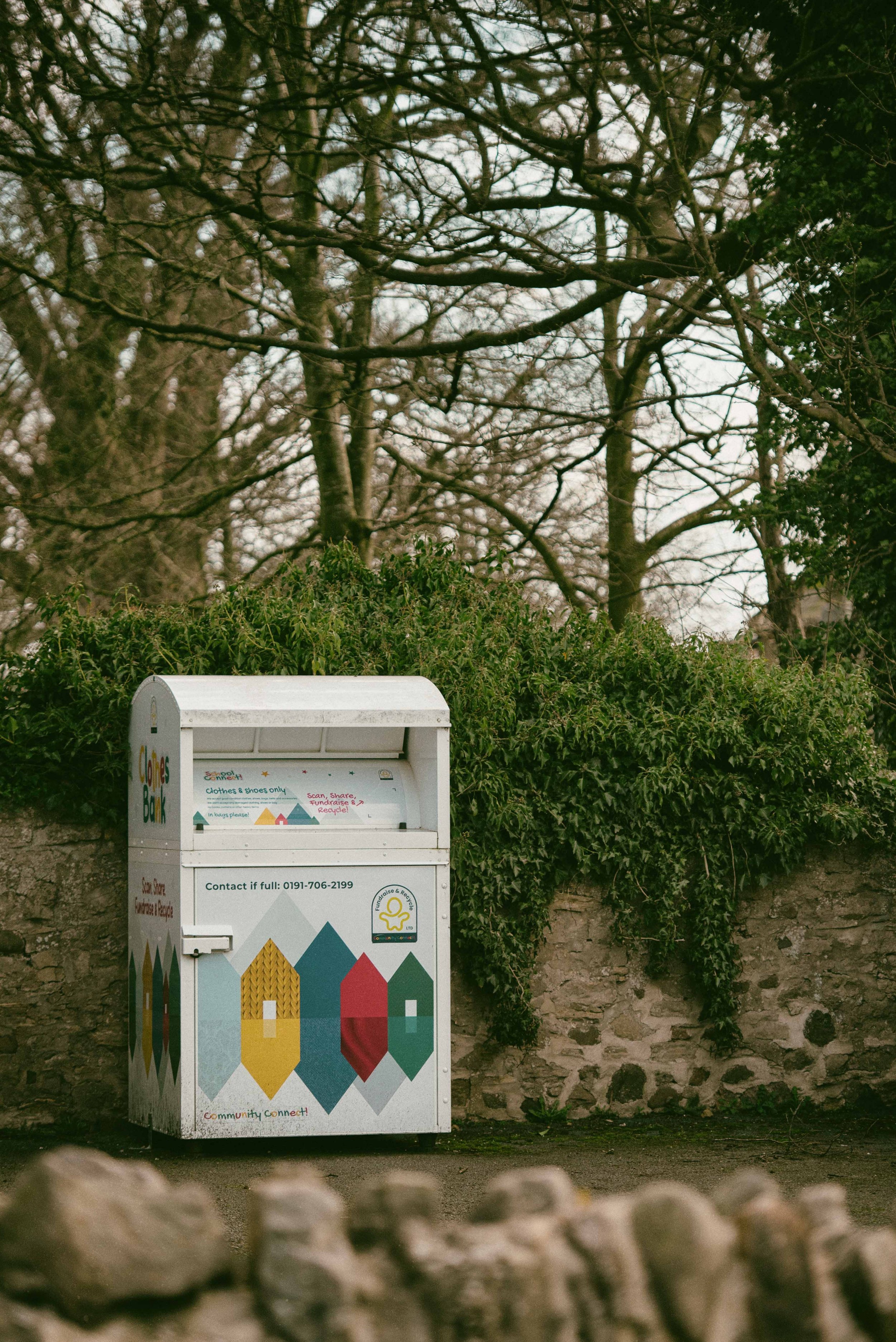 A community clothes and shoes donation bin placed against a stone wall and green bushes, with leafless trees in the background on an overcast day.