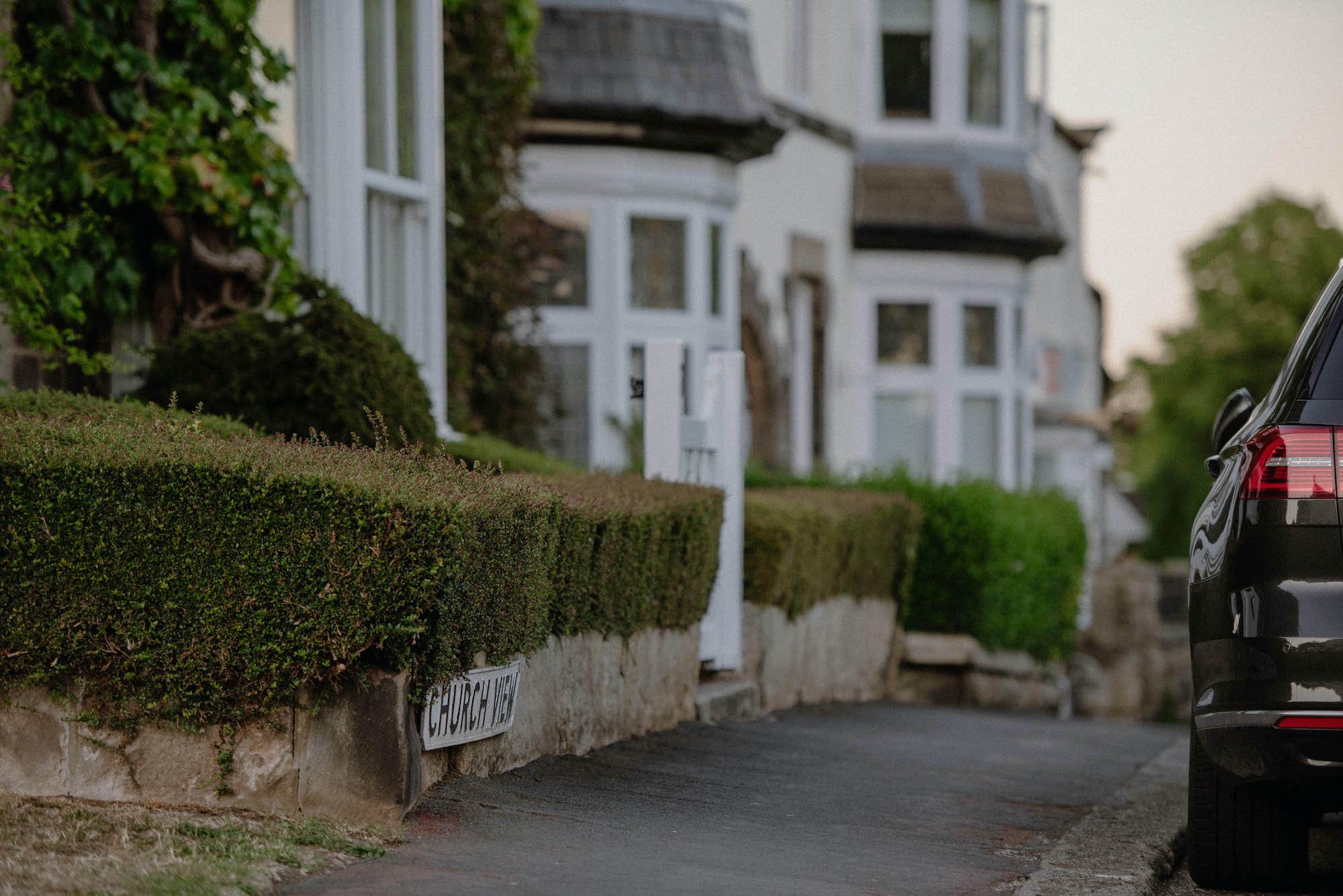 Row of white houses with a small front yard and a hedge, parked black car in the foreground, street sign reading 'Church Hill', blurred background, residential neighborhood, overcast sky.