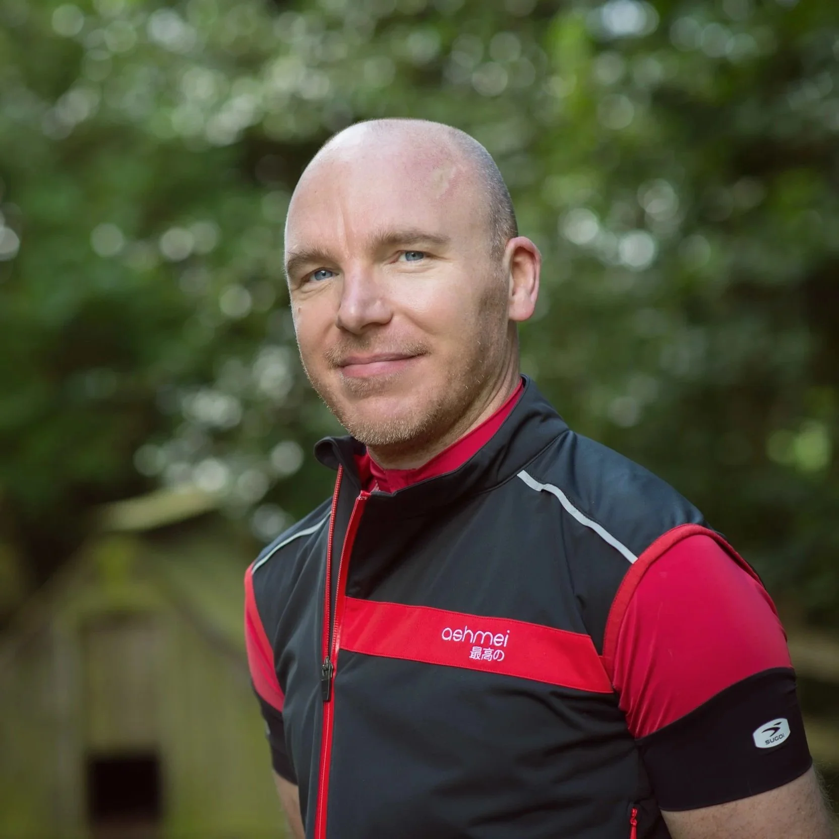 A man with a shaved head and light stubble wearing a black and red sports vest with the brand name 'ashmei' and Japanese characters, standing outdoors with a blurred green background of trees.
