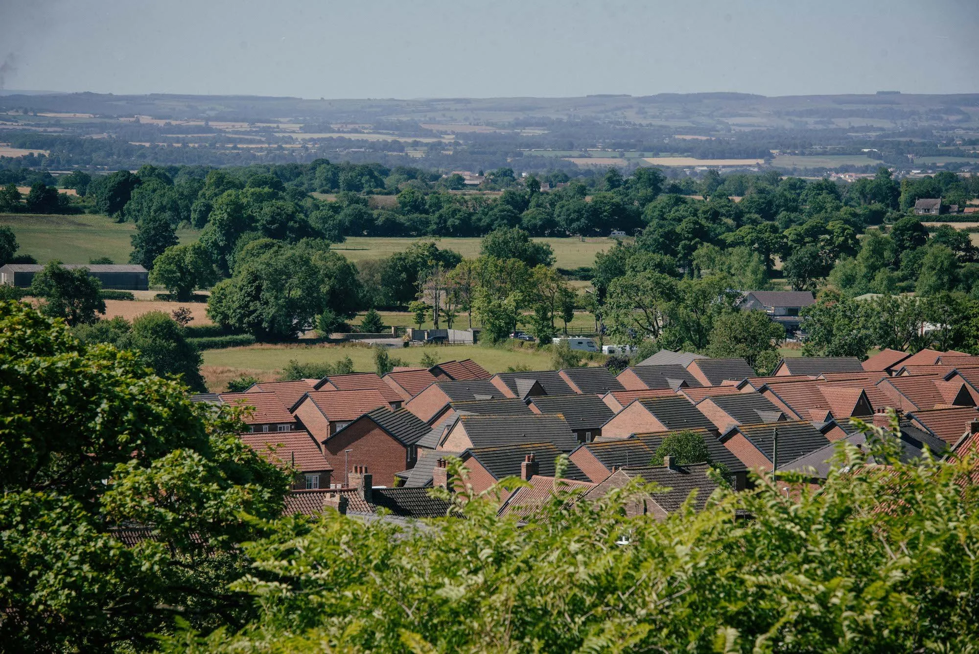 Aerial view of a residential neighborhood with row houses featuring red-tile roofs, surrounded by lush green trees, open fields, and distant rolling hills under a hazy sky.