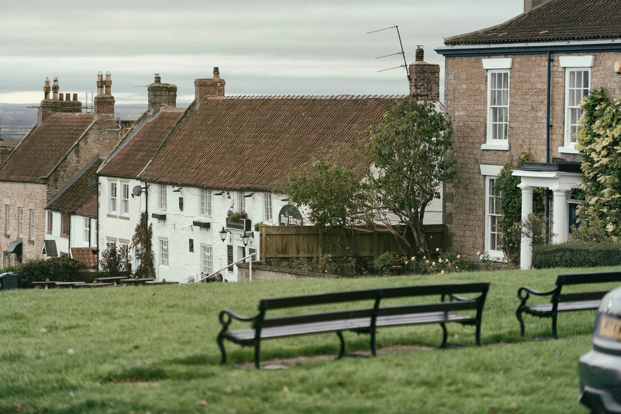 A quiet neighborhood in Heighington with stone and brick houses featuring red tiled roofs, seen from the Green with benches and a grassy area, under a cloudy sky.