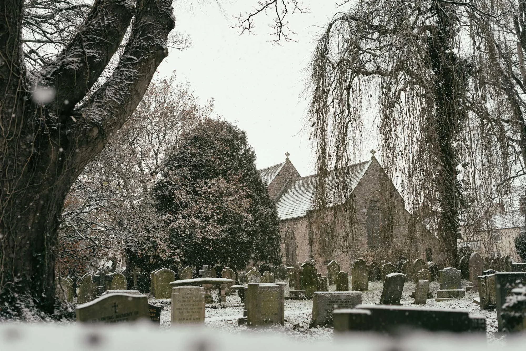 A snowy graveyard with tombstones, large trees with snow-covered branches, and a stone church in the background.