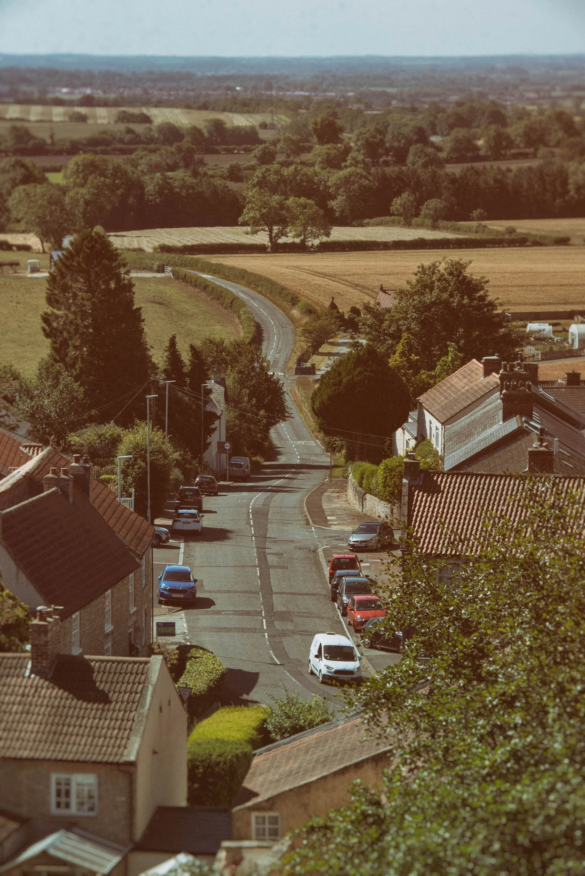 A small street with cars parked along the sides, surrounded by houses and trees, leading into a rural landscape of open fields and distant hills under a clear sky.