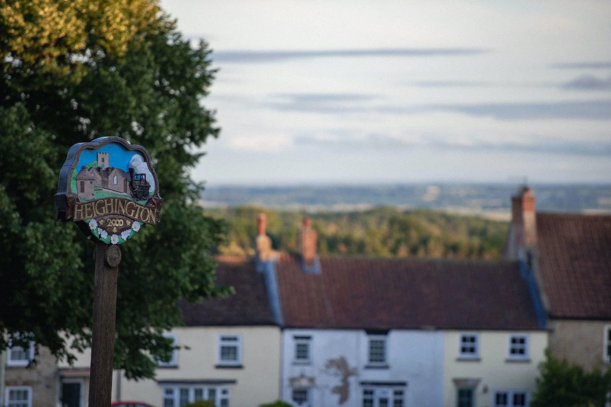 A colorful sign with a drawing of a church and the word "HEIGHINGTON" on it. The sign is mounted on a wooden post and is set against a backdrop of trees and rooftops with red brick chimneys, under a cloudy sky.