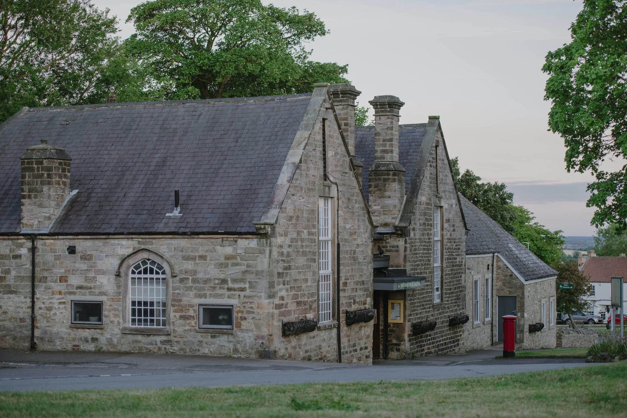 A historic stone building with large windows, a sloped slate roof, and multiple chimneys, situated in a small town with a street, a red mailbox, and green trees surrounding it.