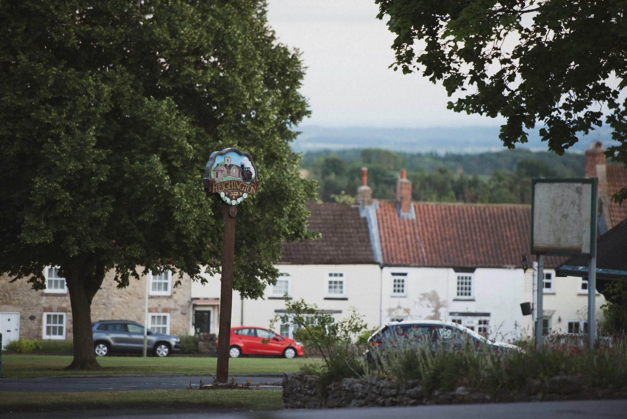 A sign with a painted scene of a church and the word "Heighington" stands next to a large leafy tree. In the background, there are houses with tiled roofs, cars parked on the street, and a golf golf backboard.