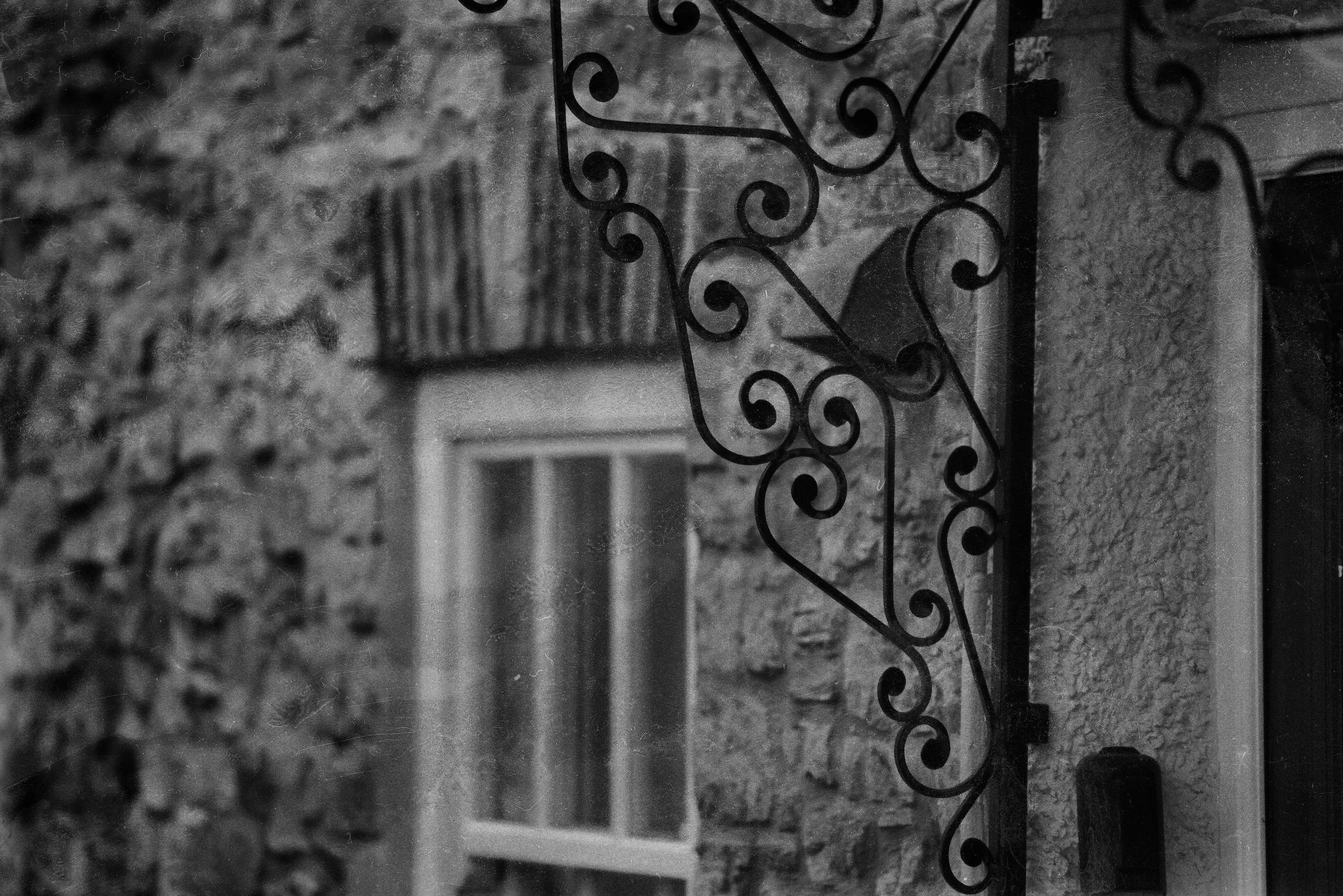 Black and white photo of ornate metalwork on a vertical surface, with a textured wall behind it and a window in the background.