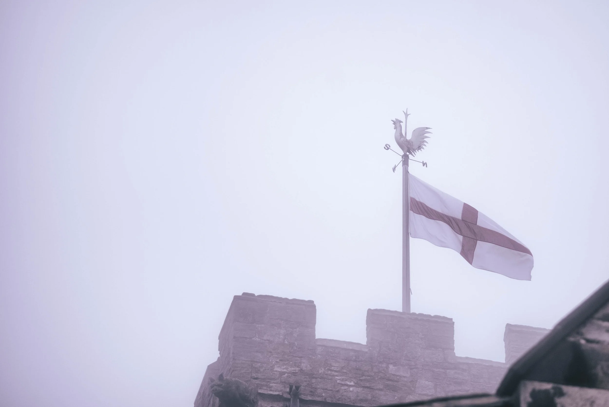 A weather vanes with a rooster design atop a stone building, flying a flag with the cross of St. George. The sky is foggy or overcast.