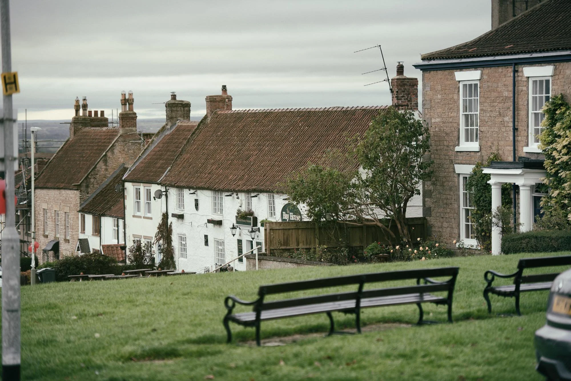 A quiet neighborhood in Heighington with stone and brick houses featuring red tiled roofs, seen from the Green with benches and a grassy area, under a cloudy sky.