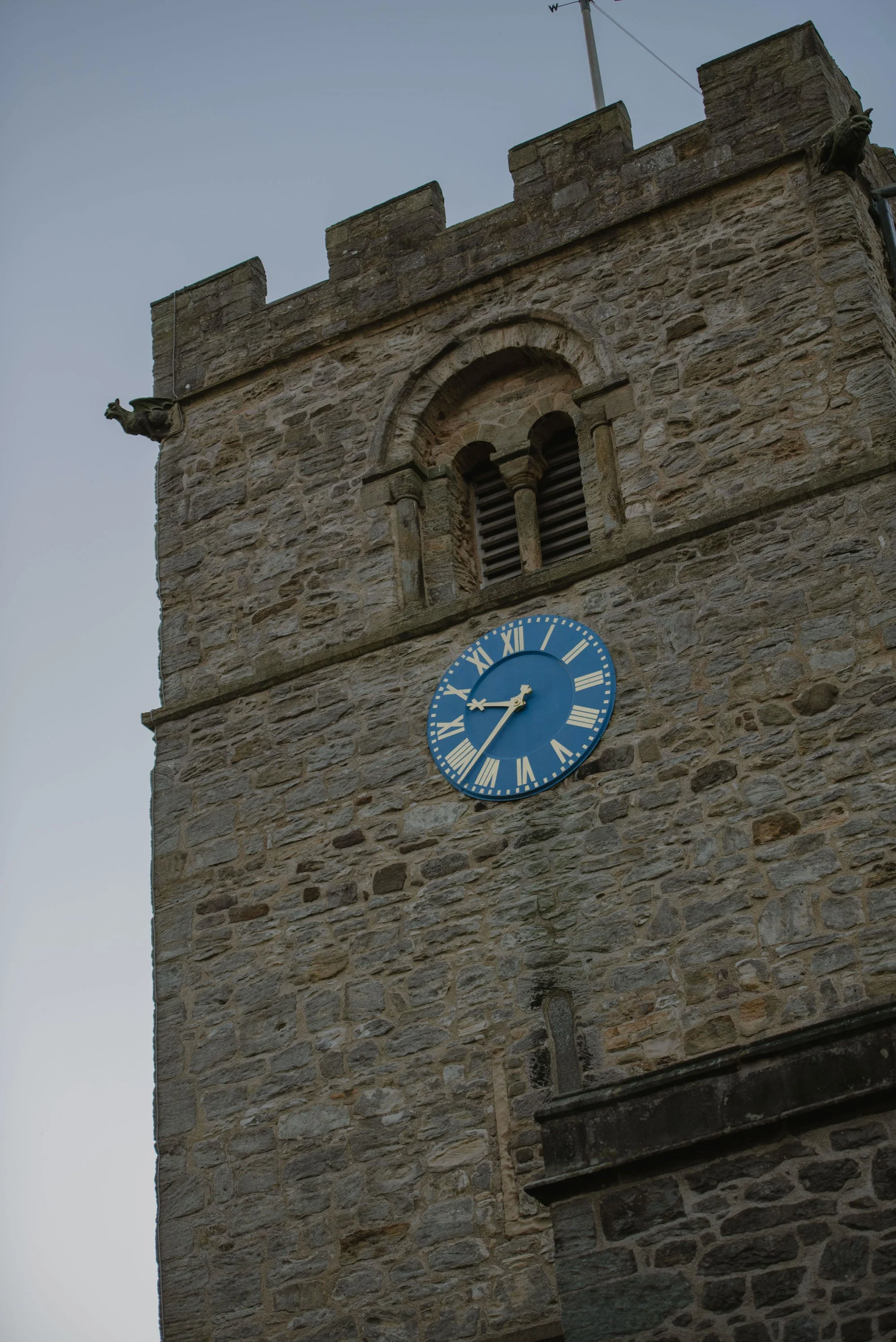 Close-up of a stone clock tower with a blue clock face, Roman numerals, and gold hands, featuring two small dragon sculptures on either side of the upper part of the tower. The tower has a small arched window with wooden slats above the clock, and an