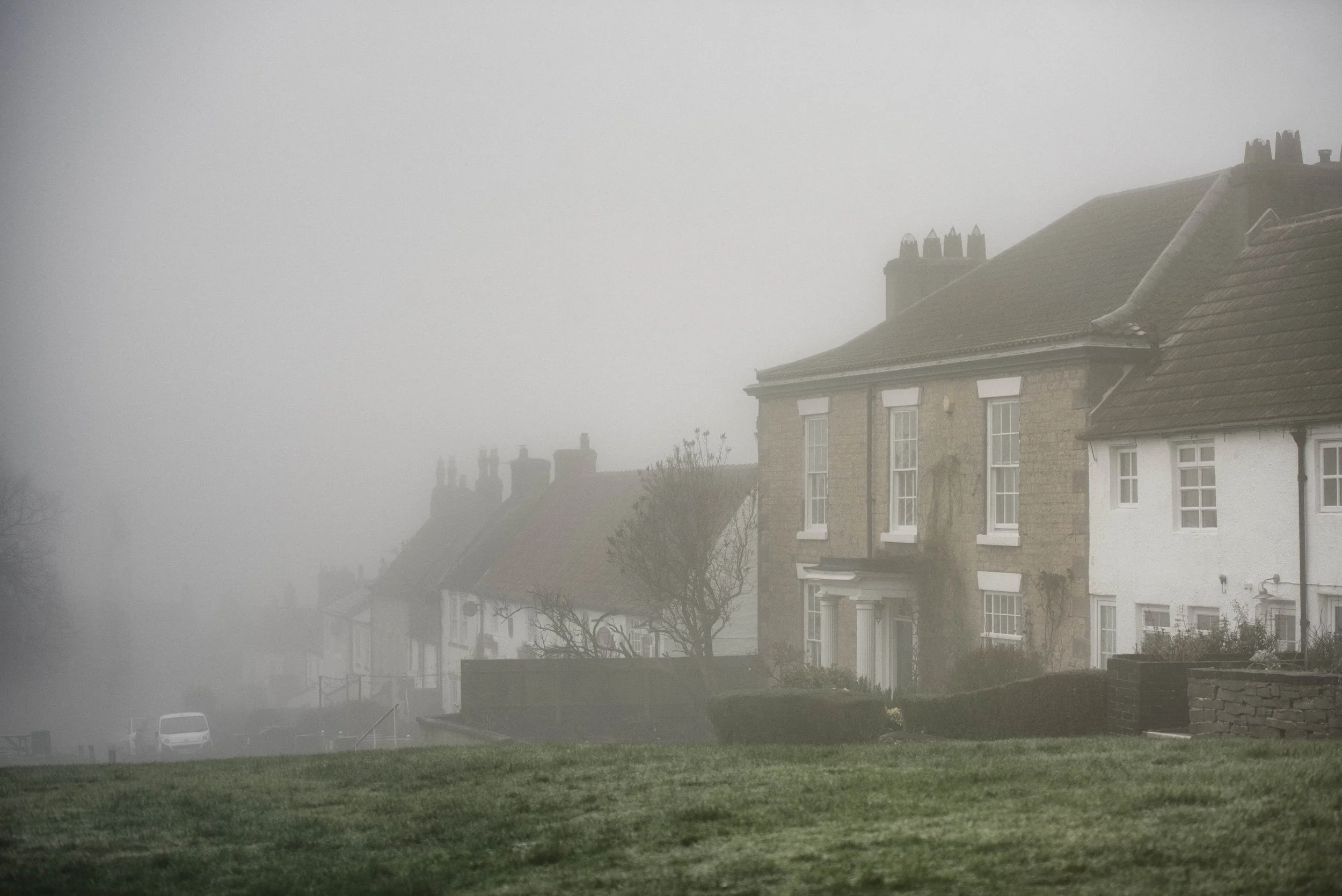 Foggy residential street with older brick and white houses, some with pitched roofs and chimneys, trees and parked cars, in a foggy atmosphere.