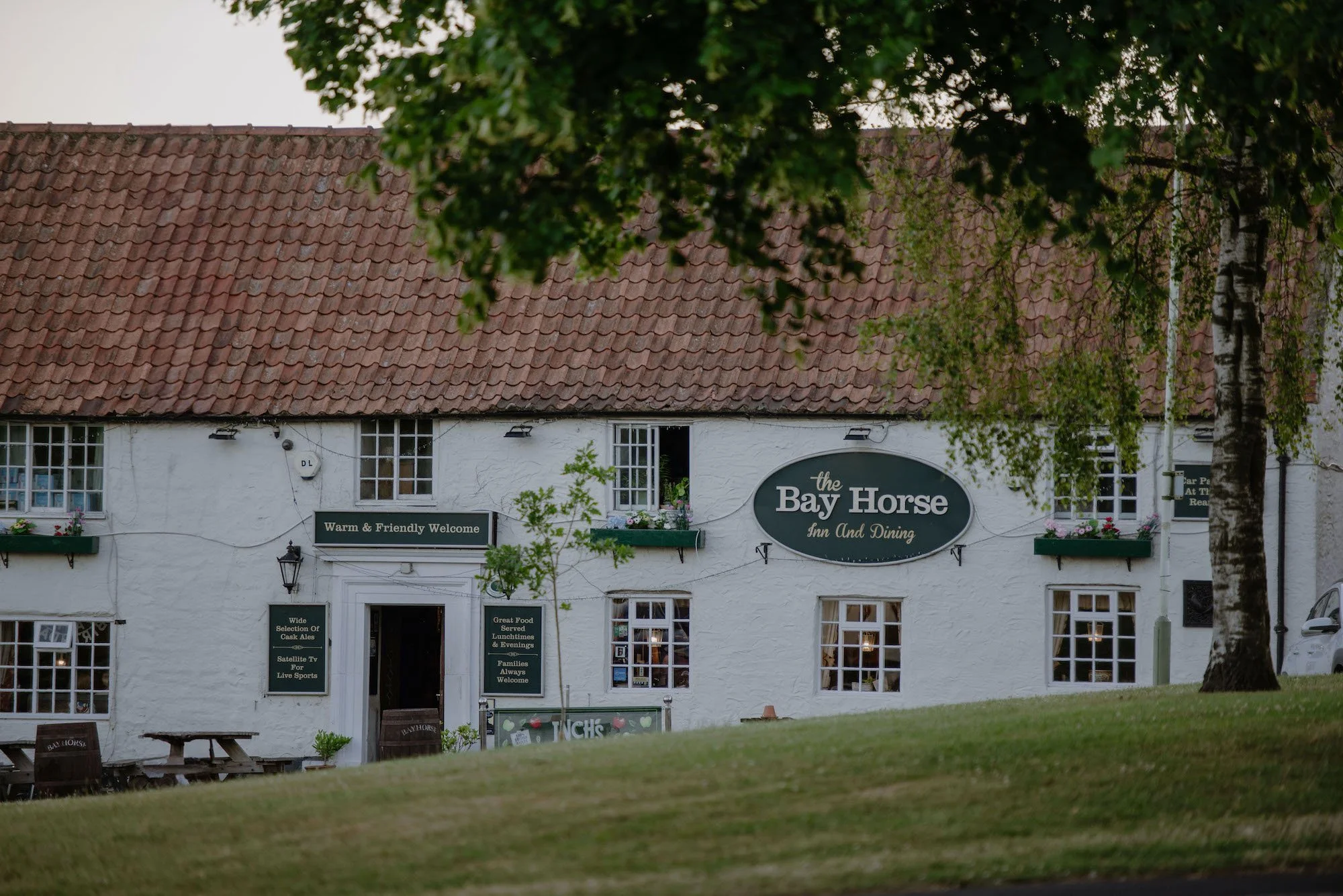 White building with a red tile roof, green trees, and a sign for "the Bay Horse Inn and Dining". The building has multiple windows with flower boxes and several signs advertising food, drinks, and entertainment. A lawn is in the foreground.