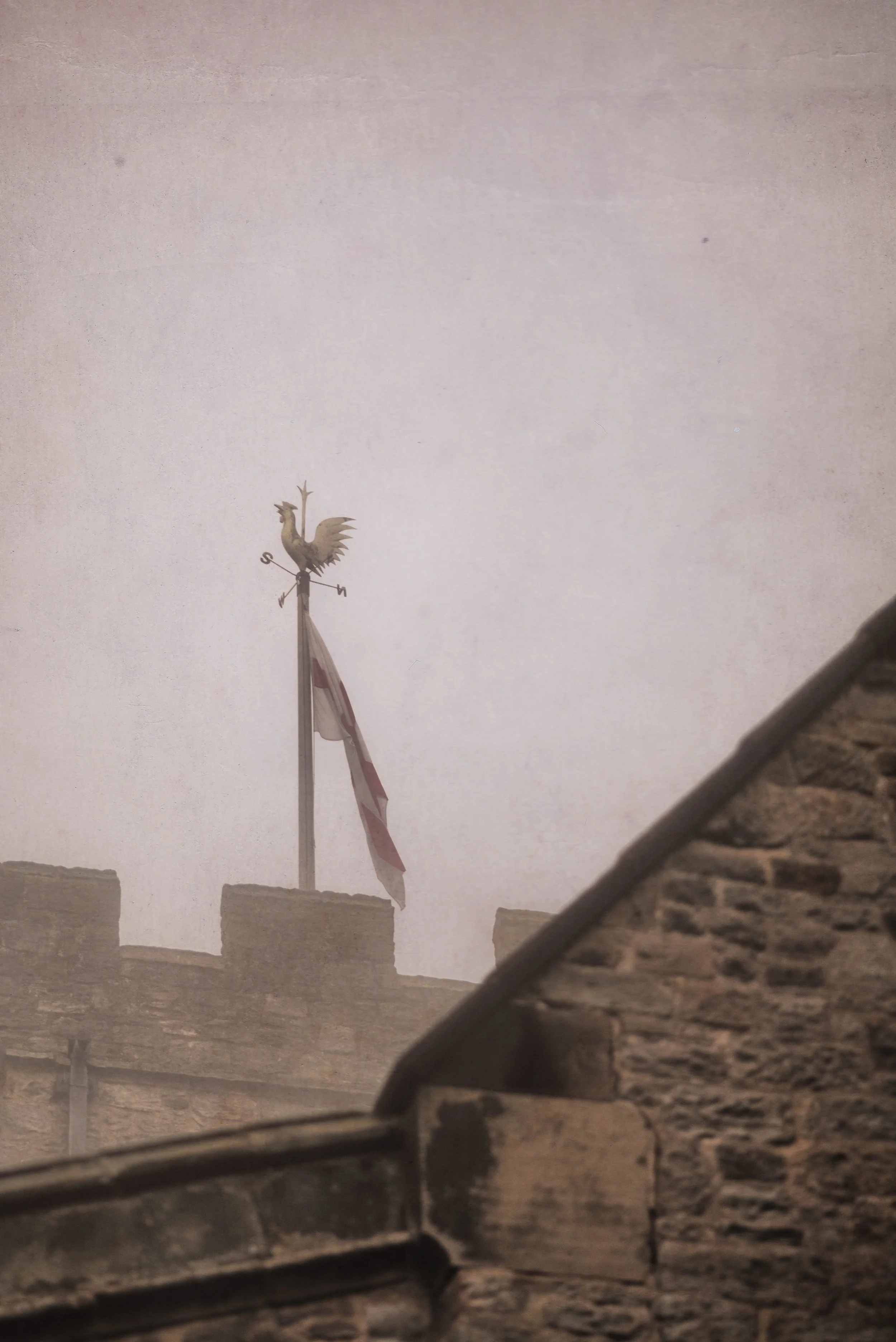 A weather vane with a rooster figure on top, mounted on a pole atop a stone building, with an American flag flying below it, against a cloudy sky background.