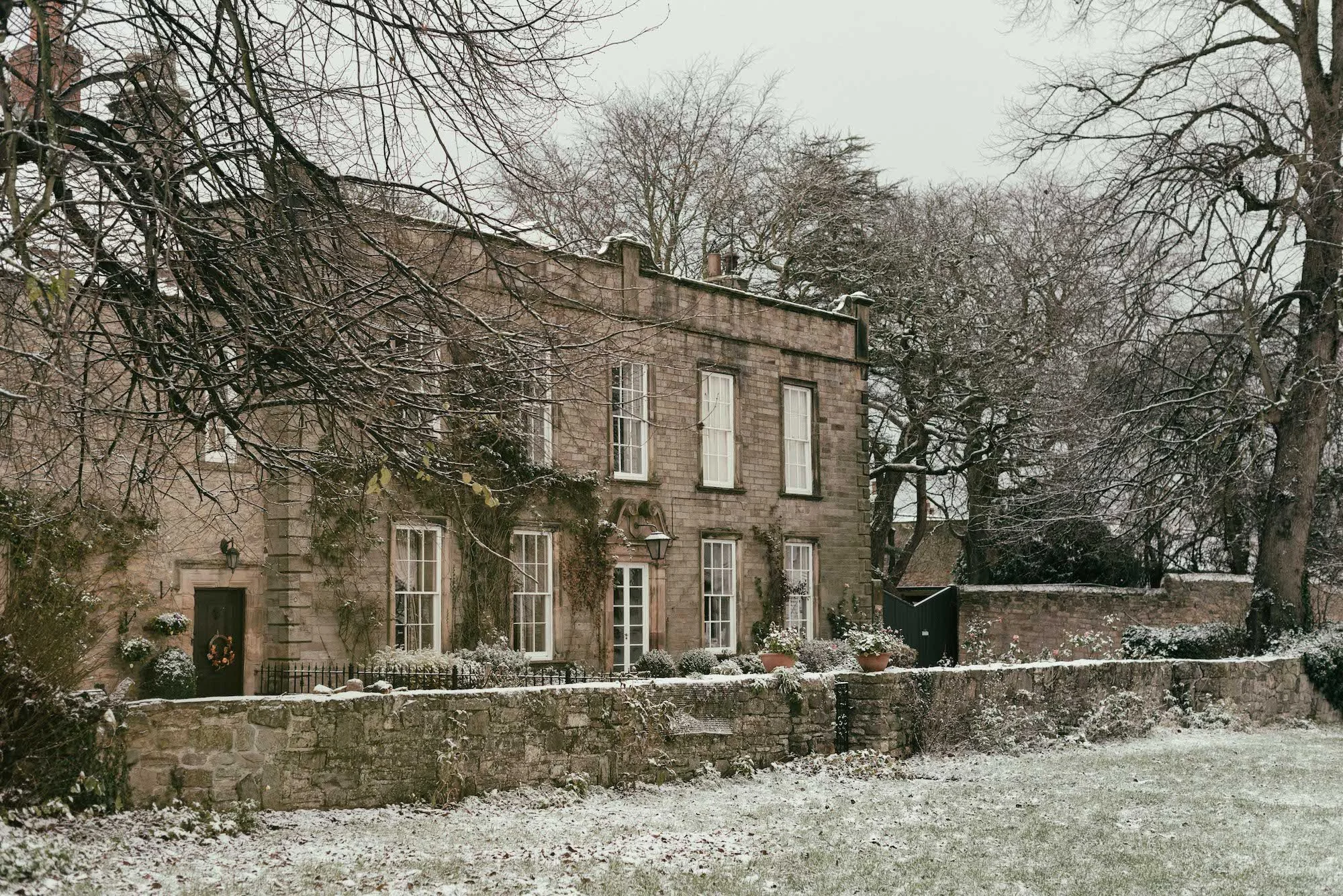 A two-story stone house with multiple windows, surrounded by a stone wall and leafless trees, with light snow covering the ground and plants.