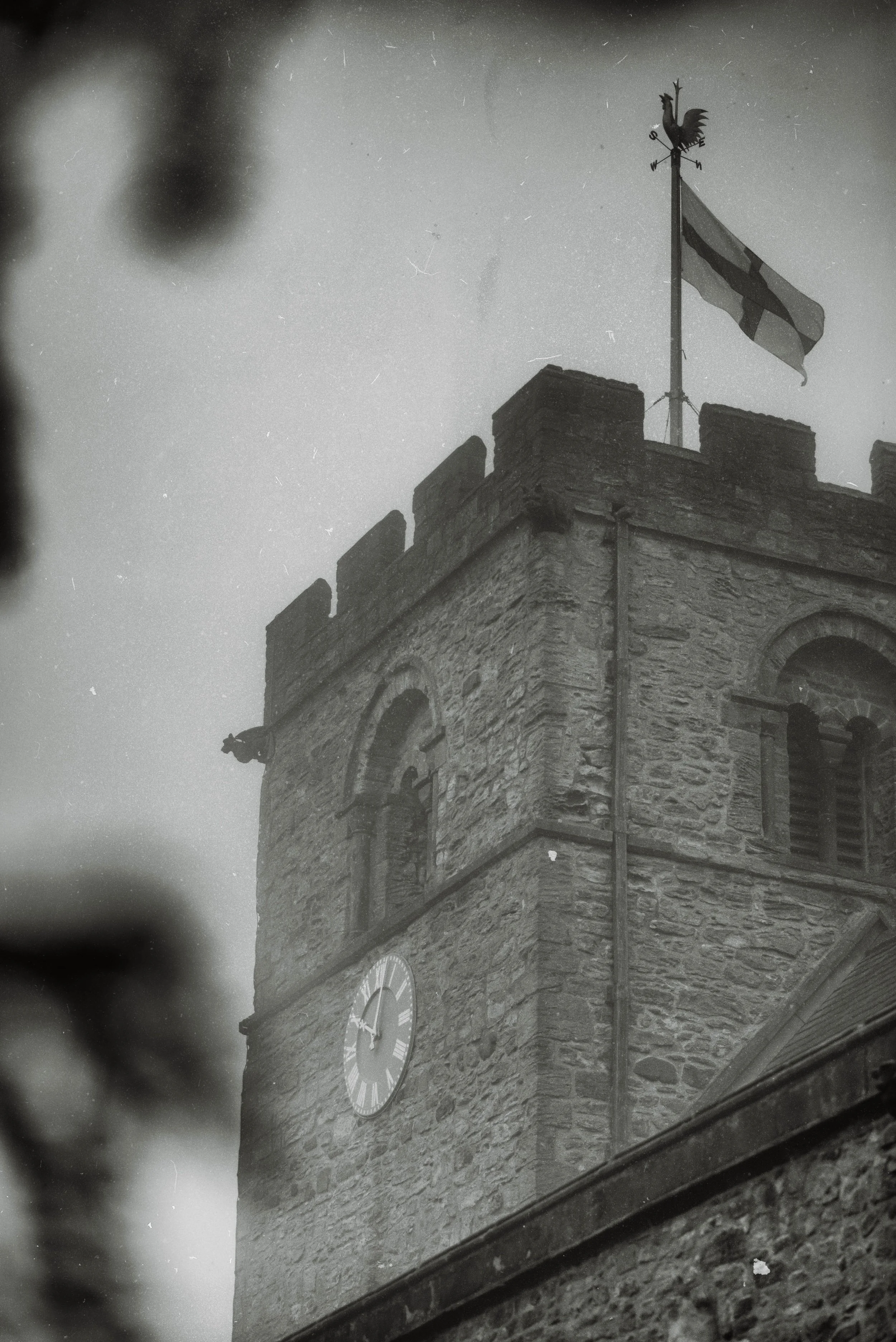 Black and white photo of a stone clock tower with a weather vane featuring a rooster on top, with a British flag flying beside it.