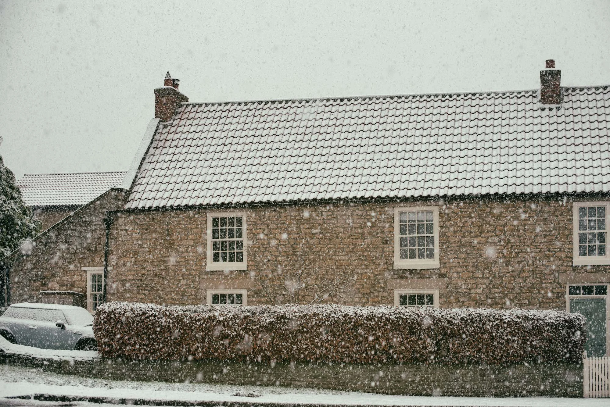 A house with a steep roof covered in snow, with multiple windows and chimneys, during a snowfall in winter.