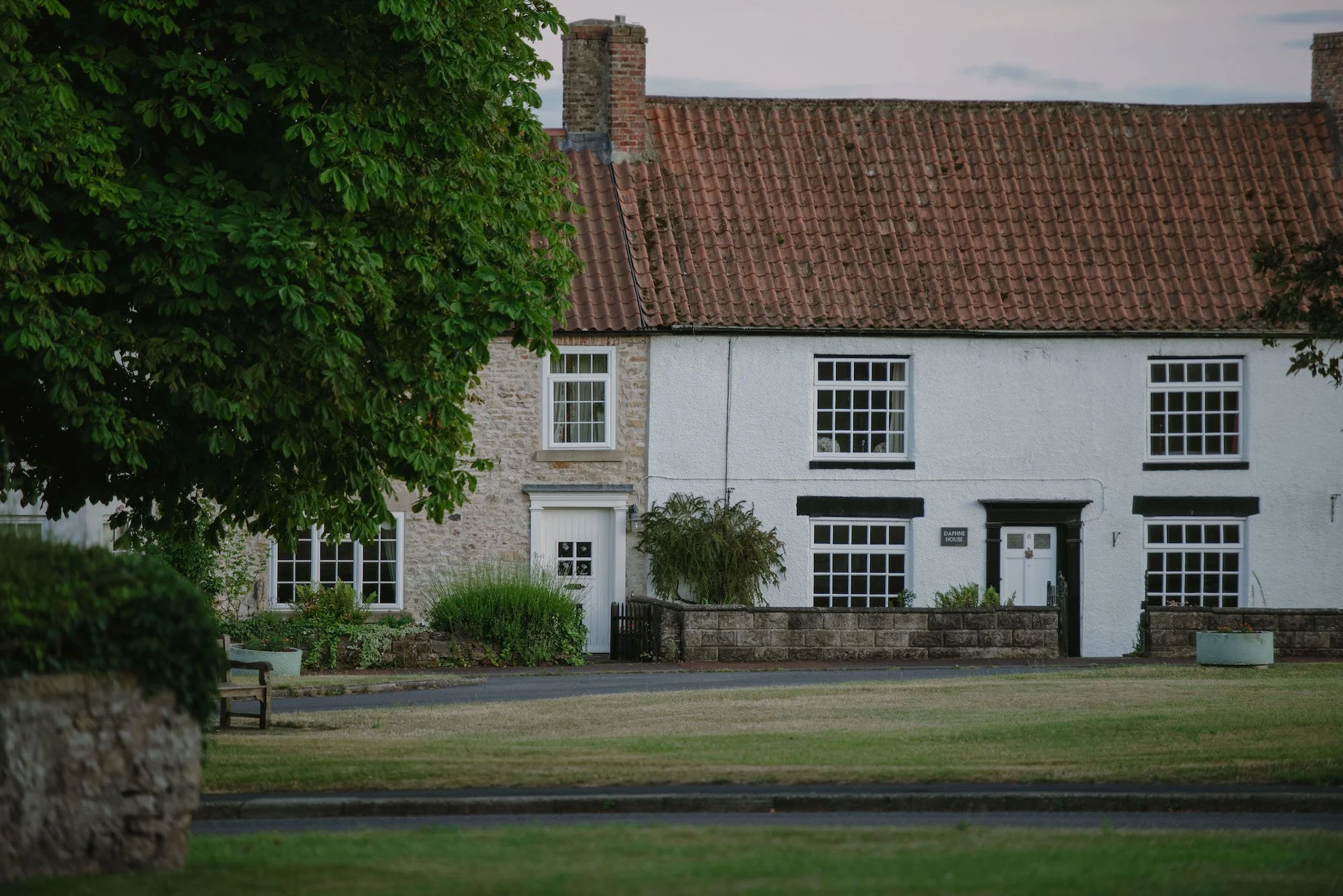 A row of white houses with brick and stone facades, red tiled roofs, and black window frames, partially obscured by a lush green tree and a stone wall in the foreground.