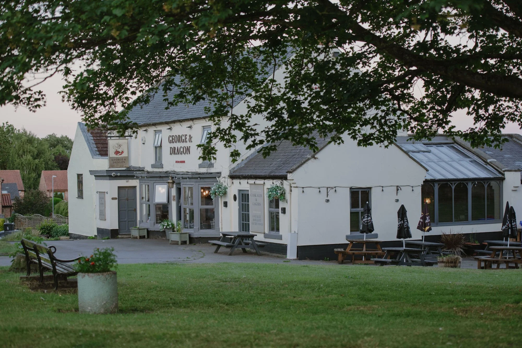 Exterior view of a white building labeled 'George & Dragon' with outdoor seating, umbrellas, greenery, and trees during daytime.