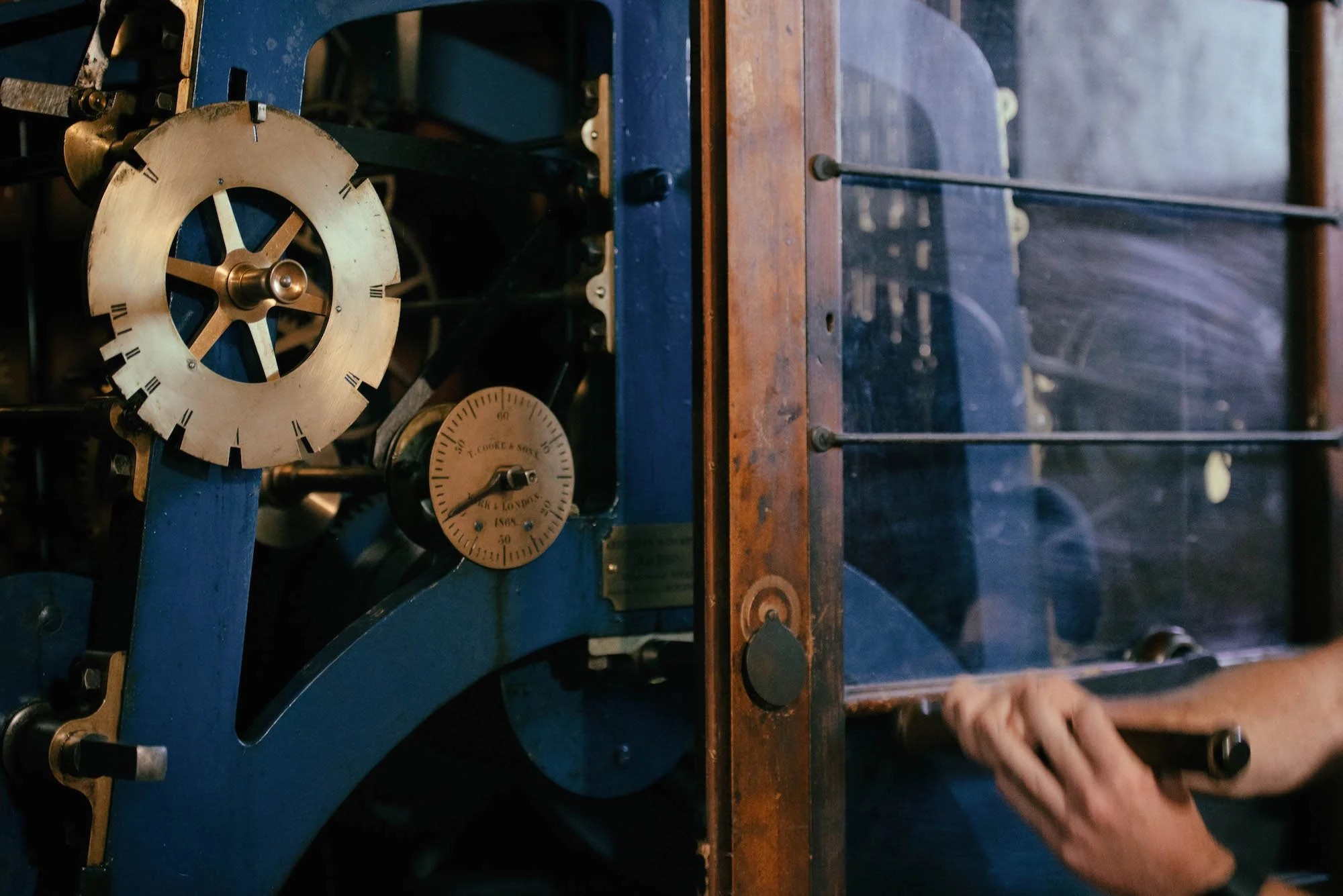 Close-up of an antique clock mechanism with a large brass gear, a pressure gauge, and various metal components, mounted on a blue and wooden frame.