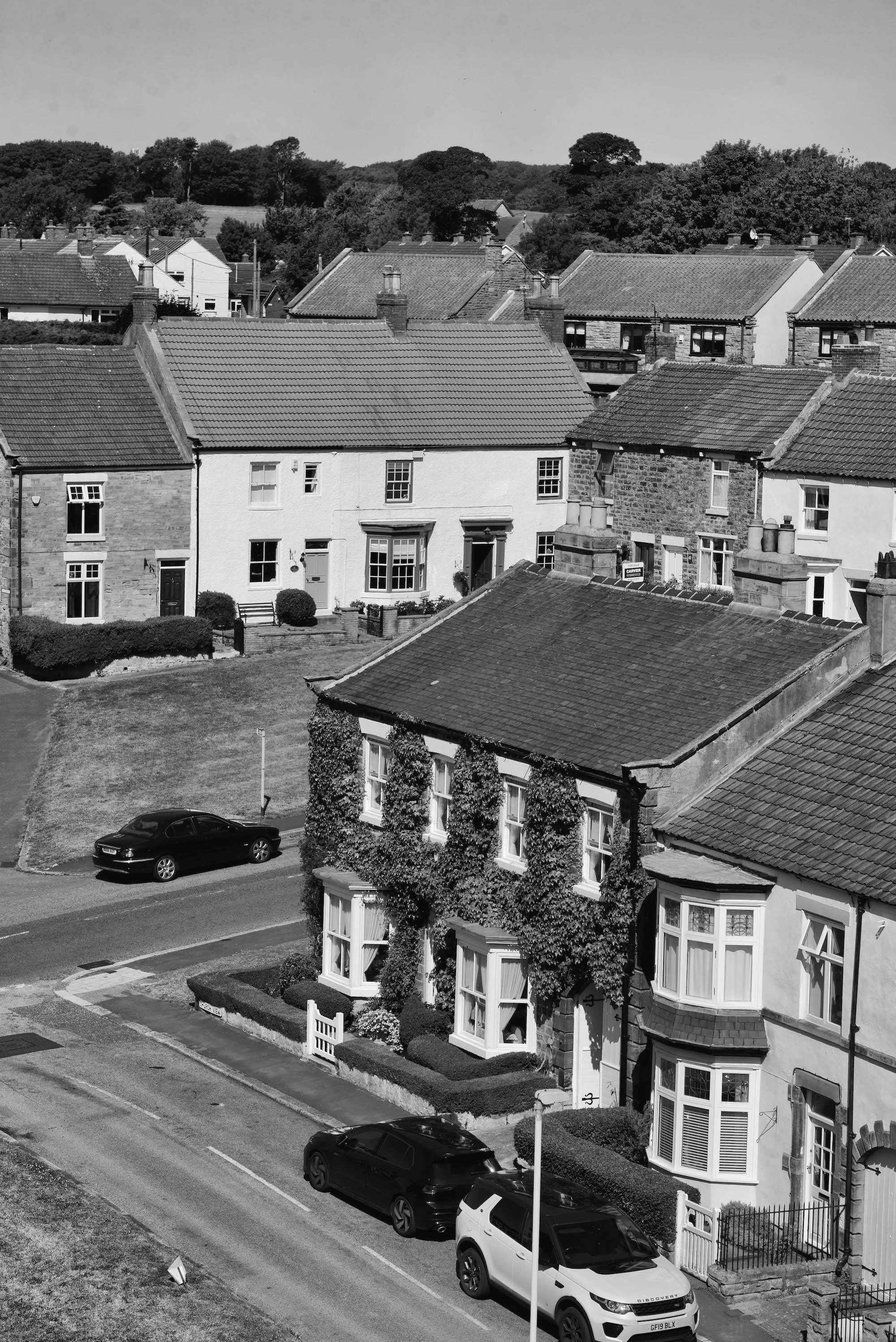 Black and white photo of a residential street with houses, cars parked along the curb, and trees in the background.