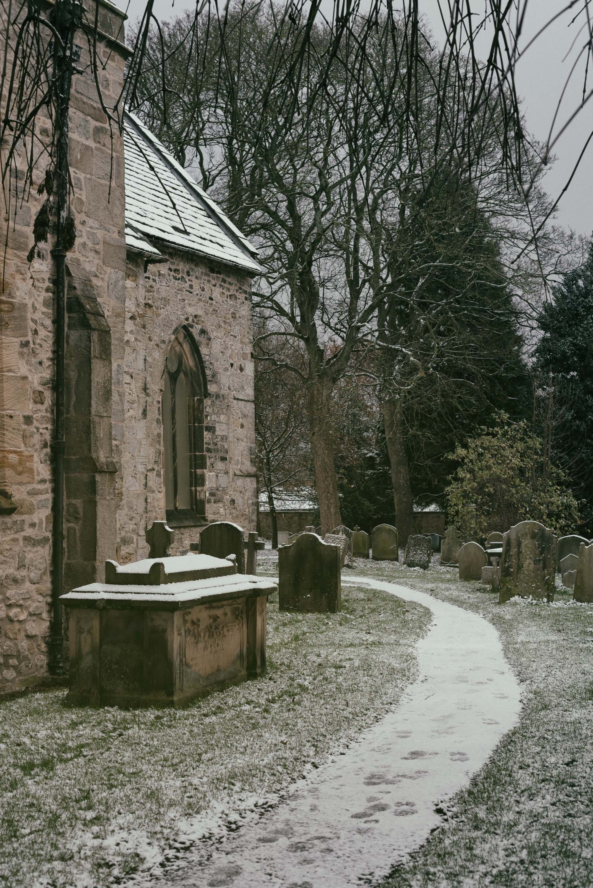 A snow-covered footpath curves past forgotten headstones and an old stone church in a snowy graveyard during winter.