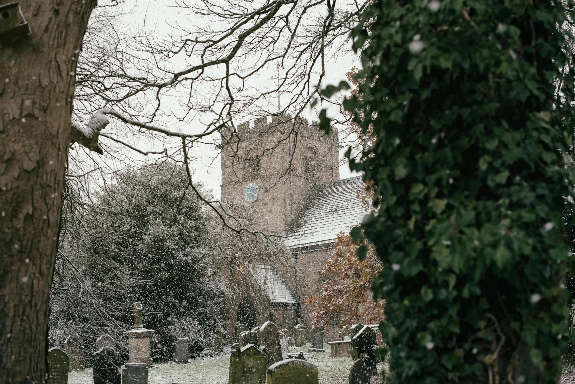 A snowy scene of a church with a clock tower, framed by a large tree on the left and a hedge on the right, with snow-covered tombstones in the churchyard.