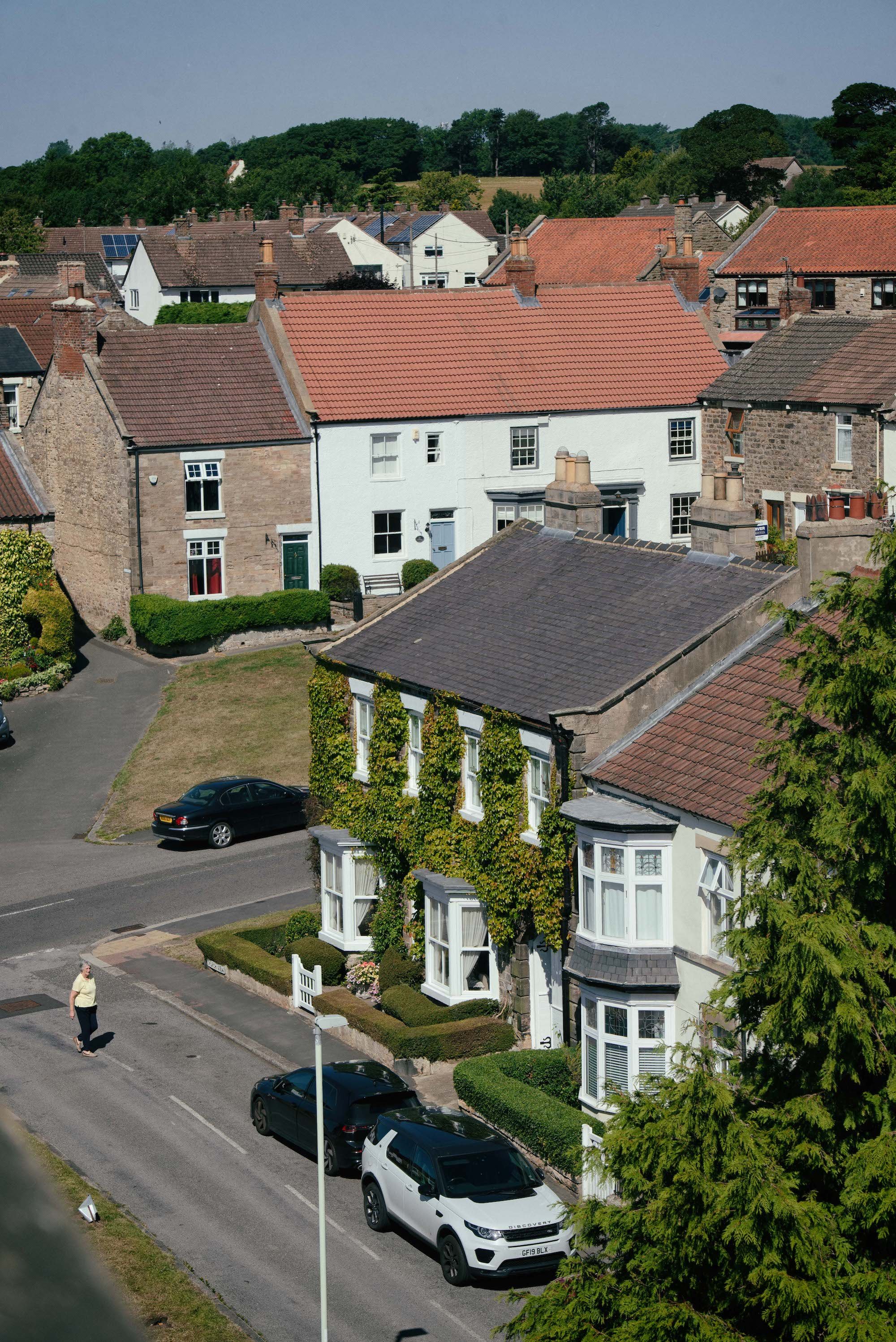 A row of houses with red and brown tiled roofs in a suburban neighborhood, some with chimneys and solar panels, lush green trees in the background, a sidewalk with a woman walking and several parked cars along the street.
