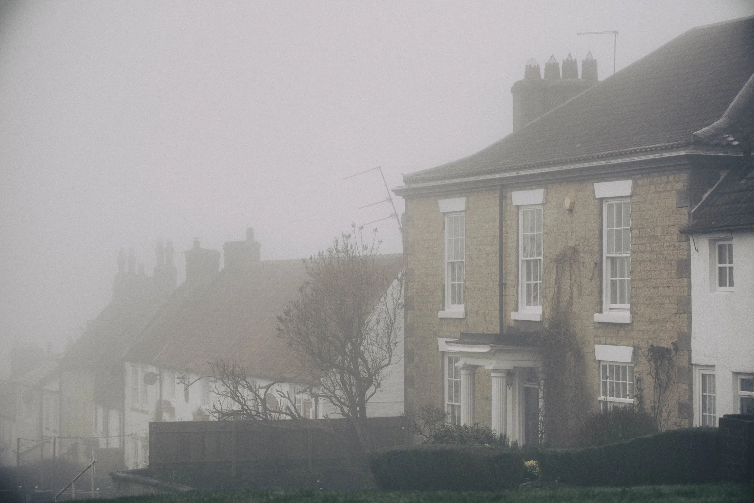A foggy daytime scene of a residential area with multi-story brick houses. The fog obscures some rooftops and chimney stacks, creating a hazy atmosphere.