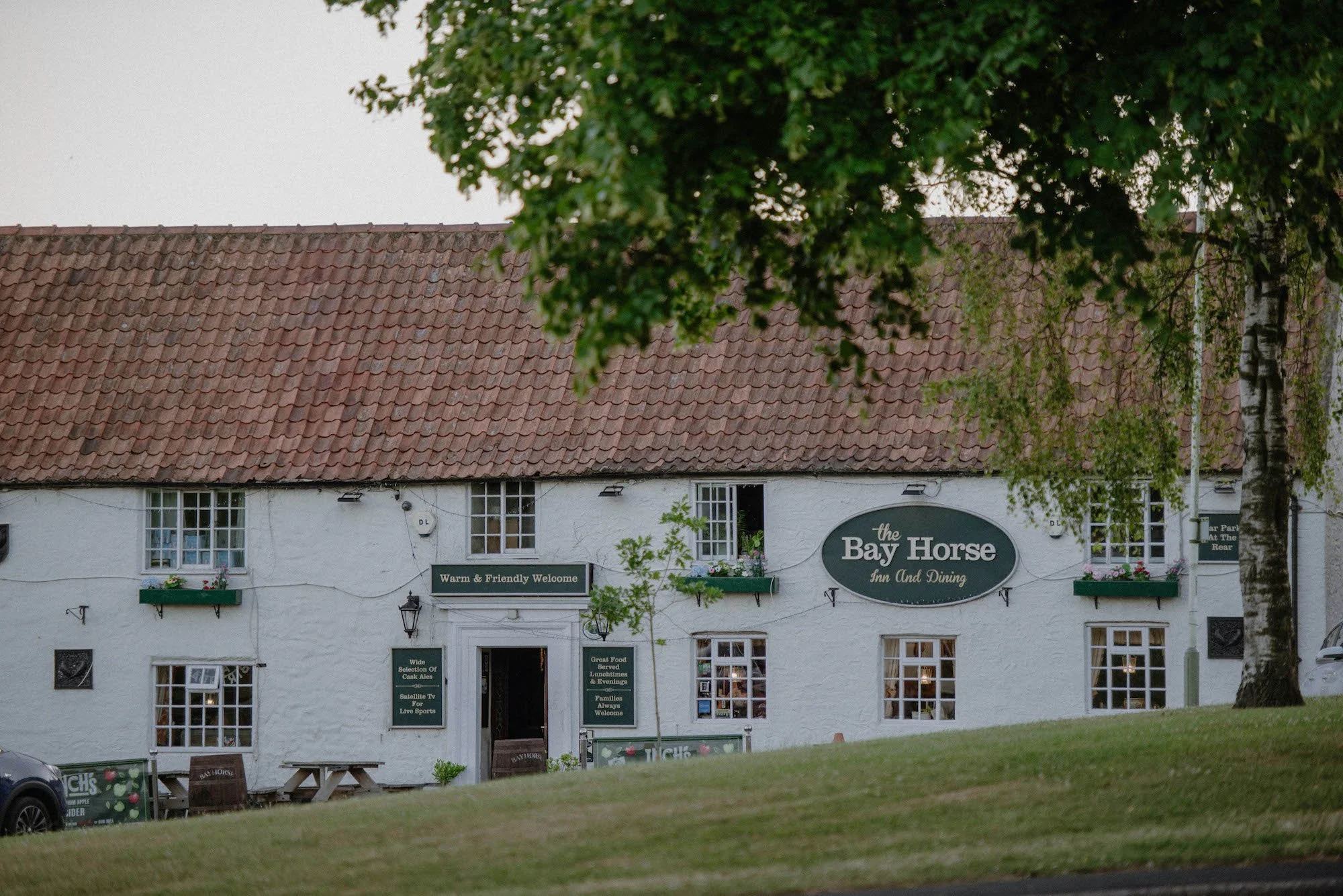 A white building with a red-tiled roof, green signs, and multiple windows. The signs read "The Bay Horse Inn and Dining," "Warm & Friendly Welcome," and other menu items. Part of a tree and a car are visible in front of the building.