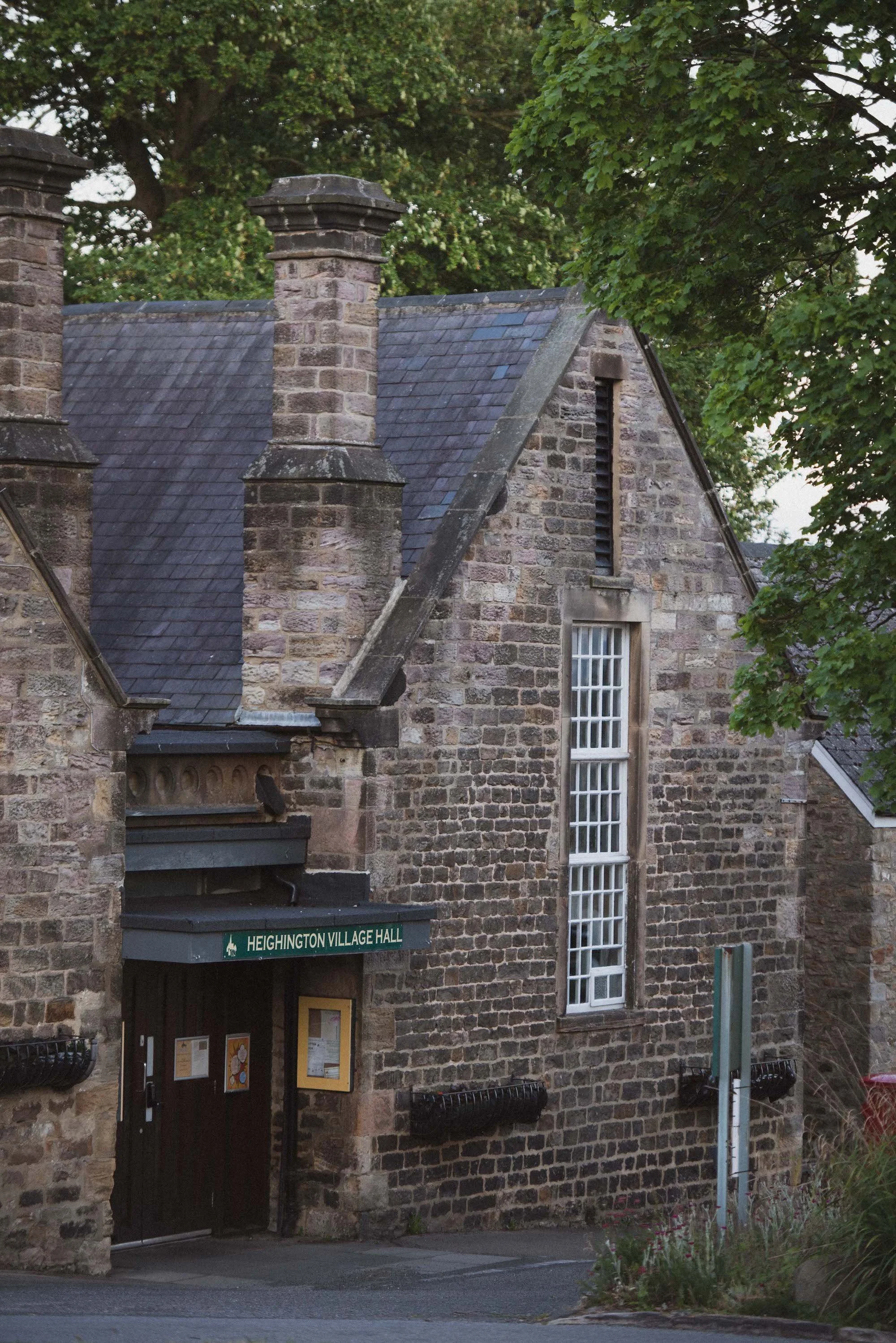 Stone building with a sign that reads 'Heighington Village Hall' above the entrance, surrounded by trees and outdoor plants.