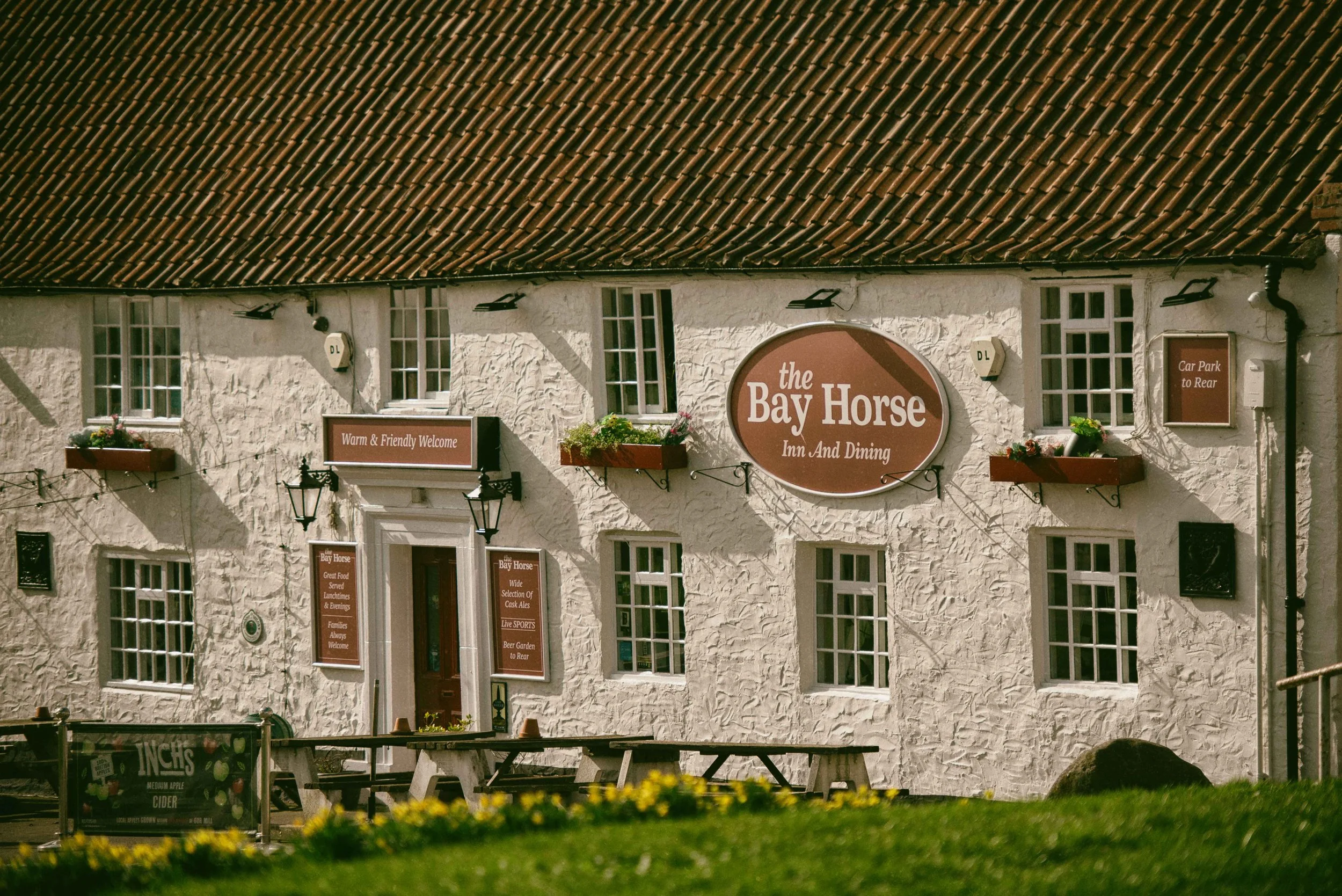 White textured building with multiple windows and signs indicating it is a pub called 'the Bay Horse Inn And Dining'. There are flower boxes outside, and signs advertising food and drinks. The roof is made of brown tiles, and there are outdoor tables