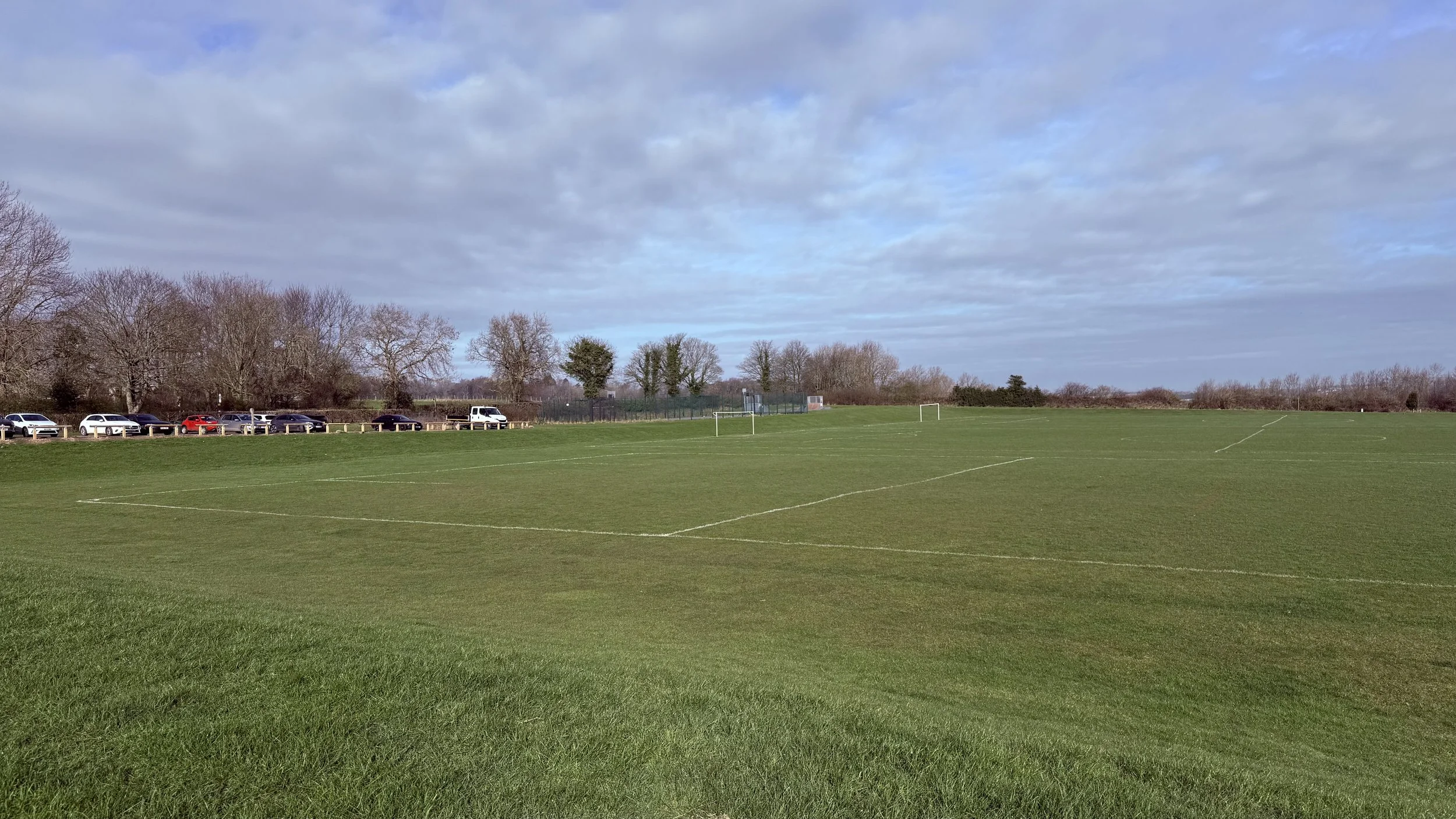 A grassy sports field with goal posts, surrounded by a parking lot with cars, and a line of trees under a cloudy sky.