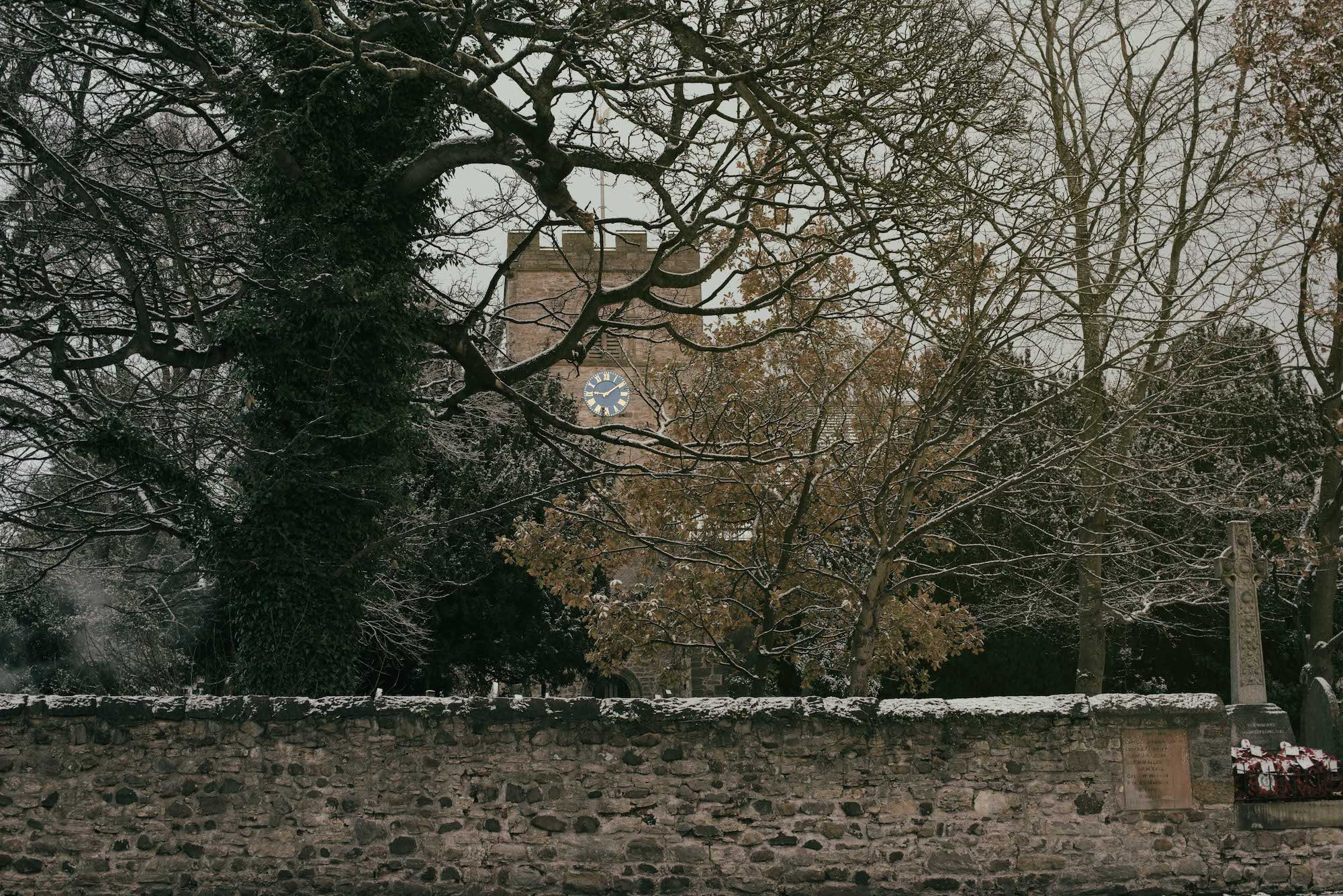 A stone wall with snow on its surface, a cast-iron fence, and leafless trees with snow on their branches. A clock tower is partially visible in the background.