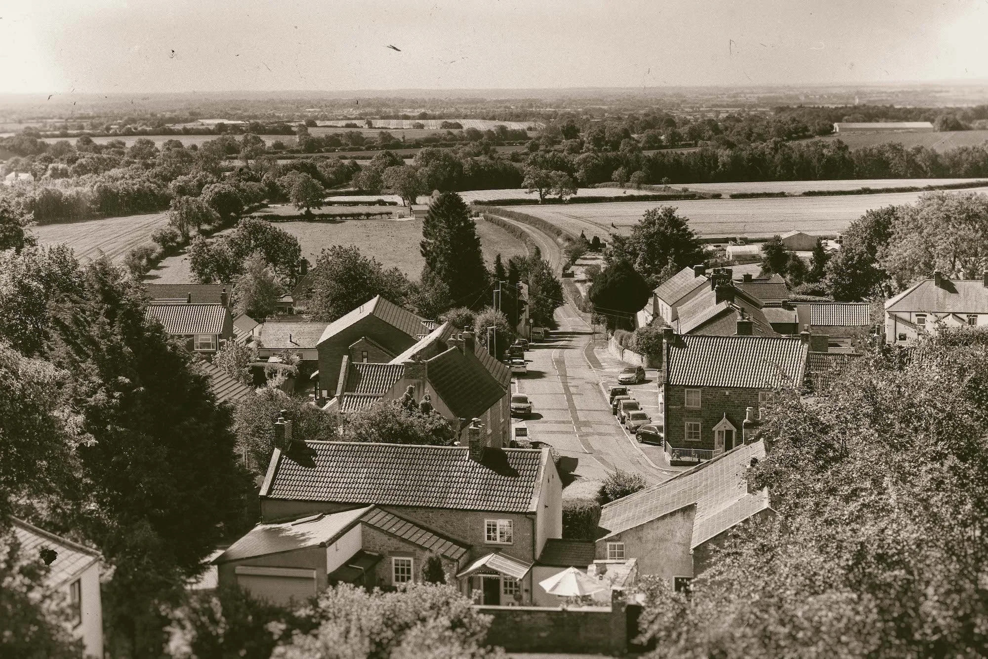 A black and white aerial view of a country village with houses, trees, and open fields, extending to the horizon.