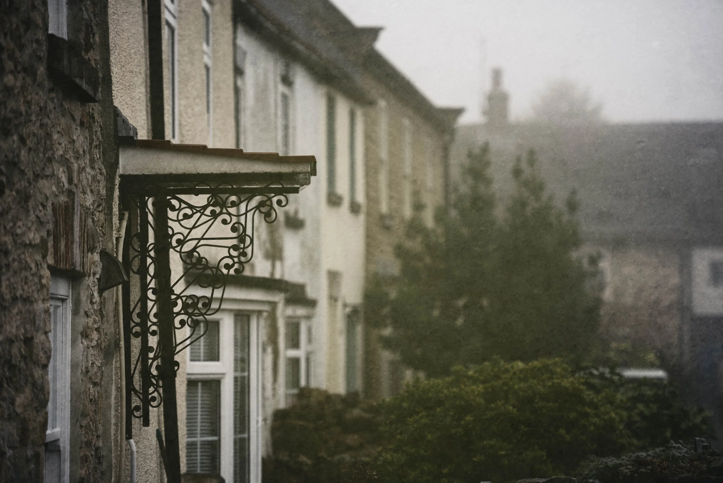 A row of residential buildings on a rainy day, with windows and a decorative black metal balcony. The sky is overcast and the scene appears misty or foggy.