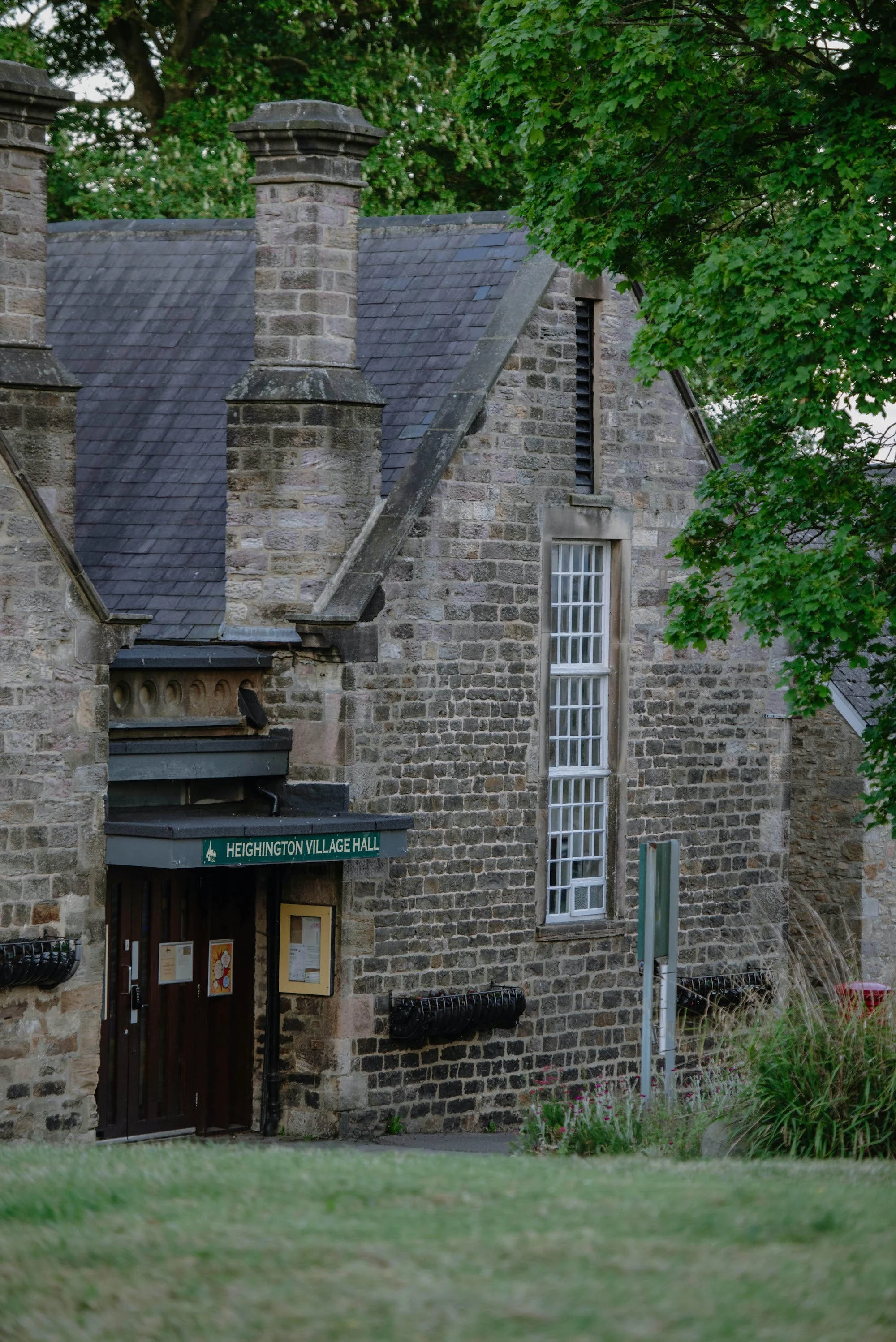 Stone building with a sign reading 'Heighington Village Hall,' surrounded by greenery and trees.