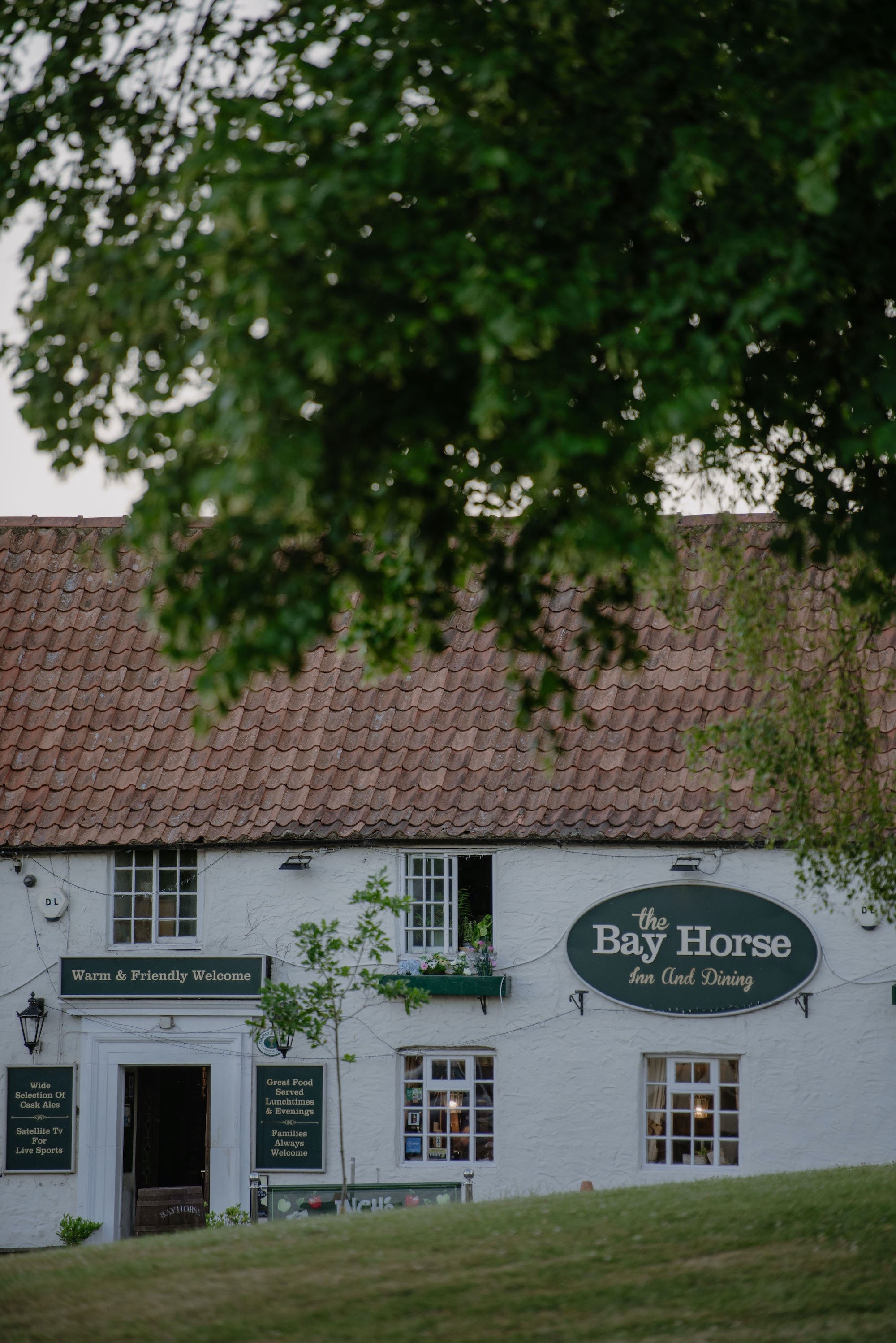 A white building with a red tiled roof and multiple windows, with signs indicating it is The Bay Horse Inn and Dining, with messages about food and entertainment. There are trees in front of the building and a lawn in the foreground.